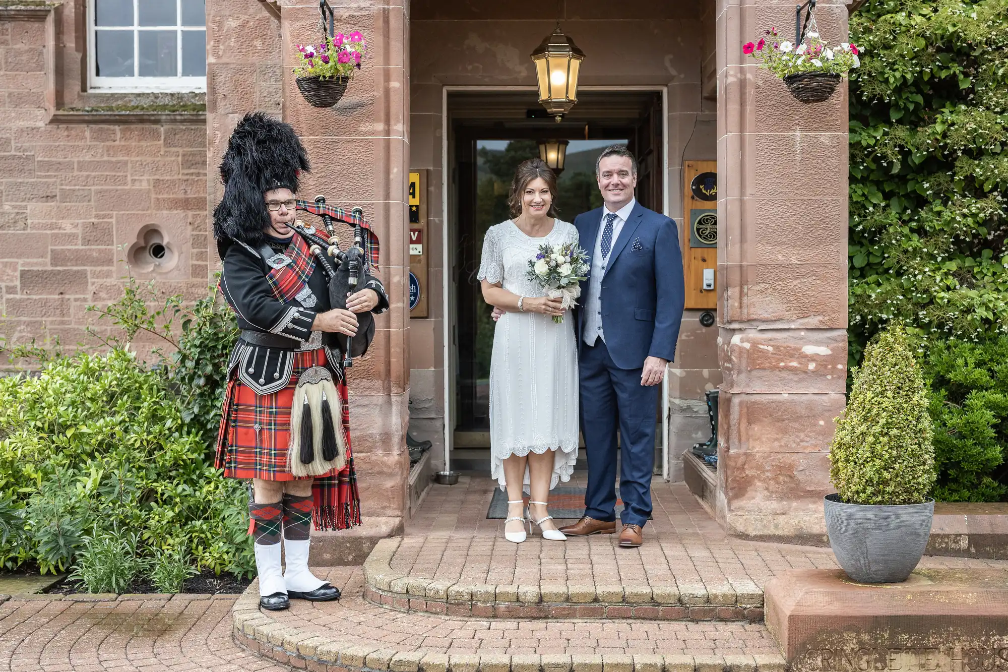 A bride in a white dress holding a bouquet stands next to a suited man at a building entrance. A bagpiper in traditional Scottish attire, including a red tartan kilt and feathered hat, plays nearby. Flowers hang on both sides of the doorway.