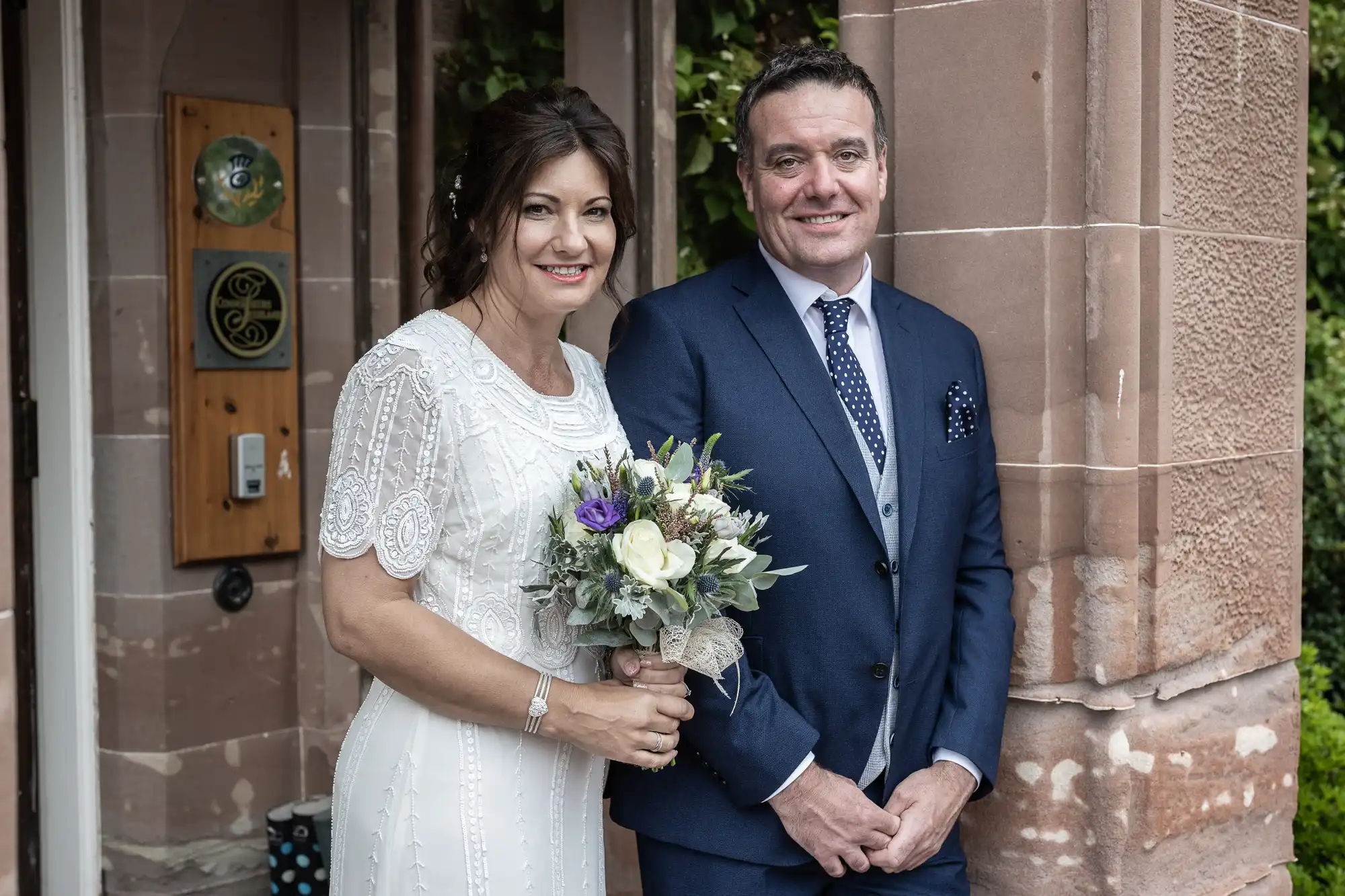 A woman in a white beaded dress holds a bouquet of white and purple flowers, standing beside a man in a blue suit with a polka dot tie. They both smile, posing in front of a stone building entrance surrounded by greenery.