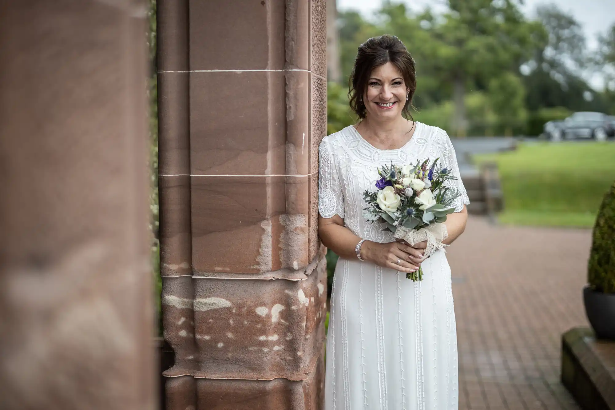 A smiling bride in a white, short-sleeved dress stands outdoors beside a stone column, holding a bouquet of white and purple flowers. Green lawns, trees, and a blurred car are visible in the background.