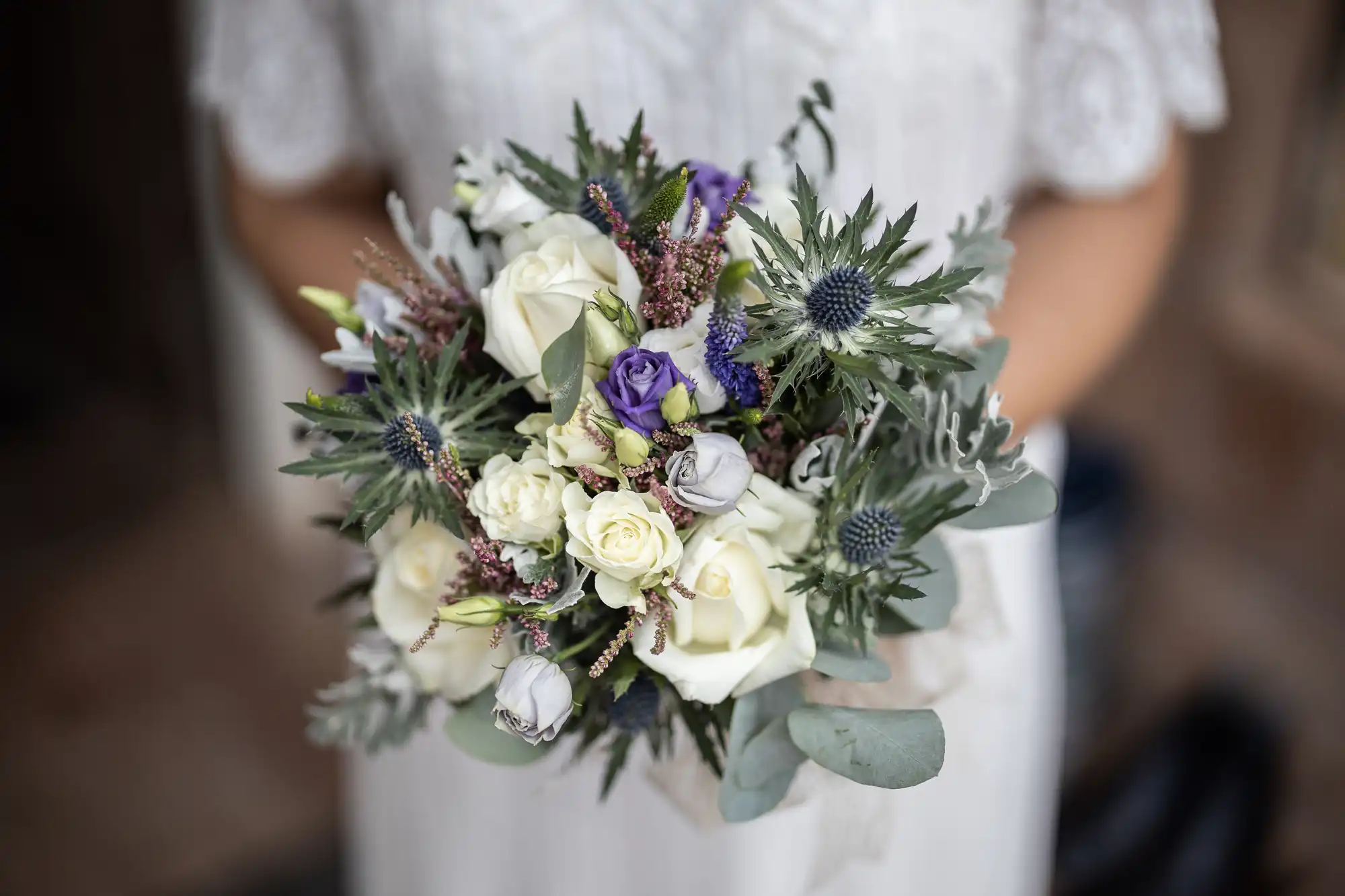 A person in a white lace dress holds a bouquet of white roses, purple flowers, thistles, and greenery. The image is focused on the bouquet, with the person's hands and upper dress visible but out of focus in the background.