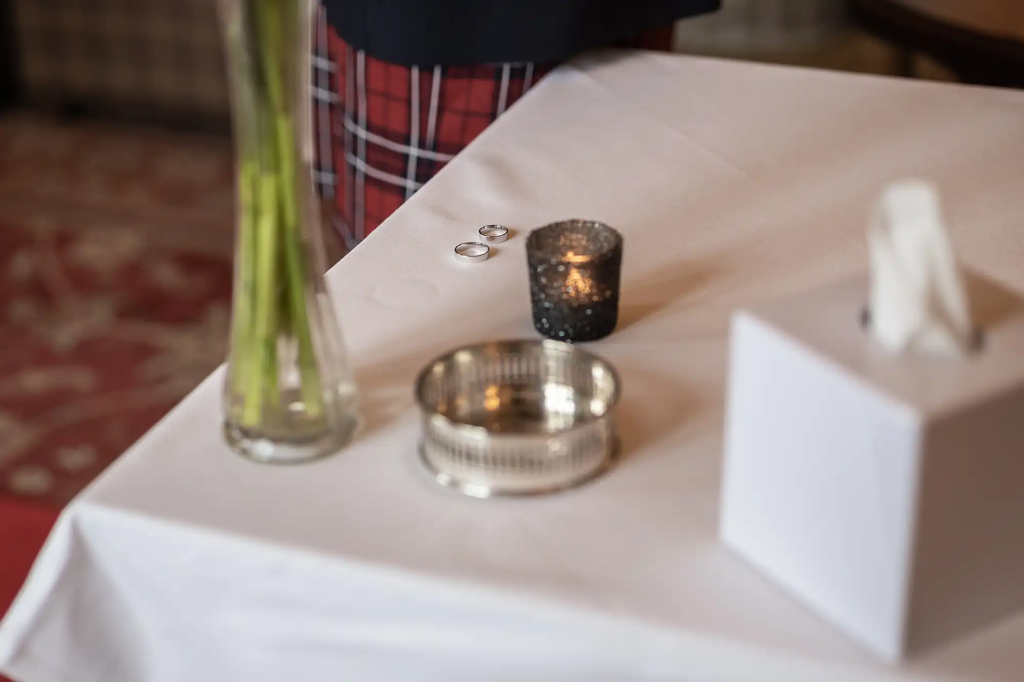 A white table holds two silver rings, a small black candle holder with a lit candle, a silver tray, a glass vase with green stems, and a white tissue box. Part of a person in checked trousers is visible in the background.