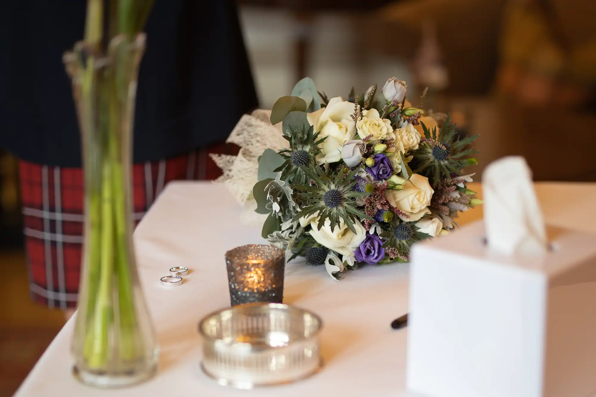 A wedding bouquet with white and purple flowers rests on a white tablecloth beside two wedding rings, a small candle holder, a silver dish, a tissue box, a pen, and a glass vase with green stems. A person in checked stands in the background.