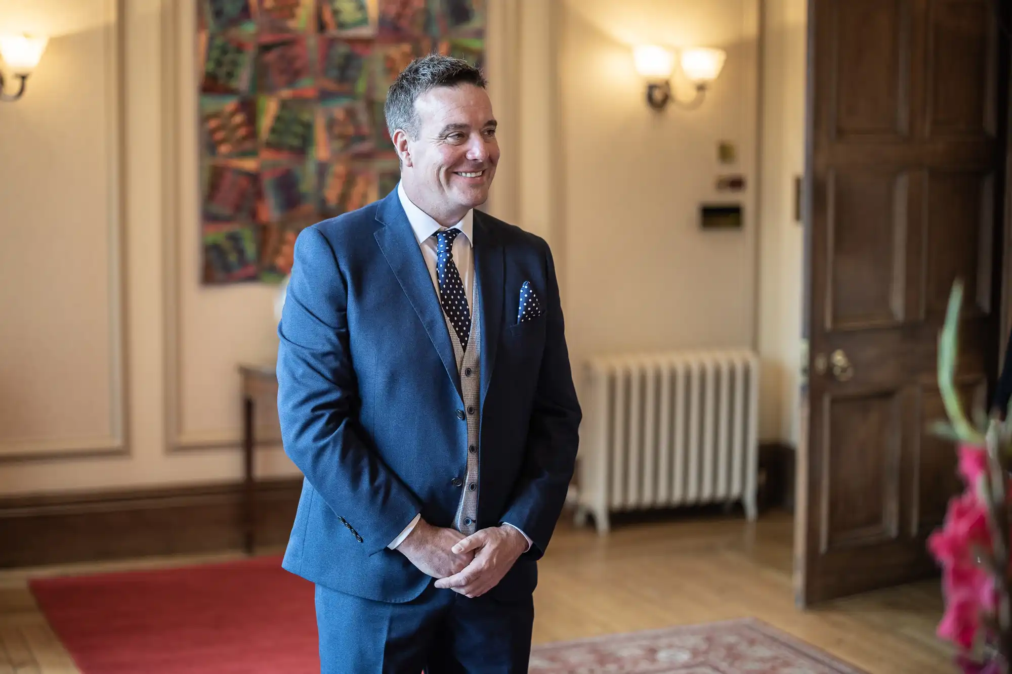 A man in a blue suit, patterned tie, and grey waistcoat stands smiling in an elegant room with wooden floors, rugs, wall art, and a radiator in the background. He holds his hands together in front of him, looking relaxed and happy.