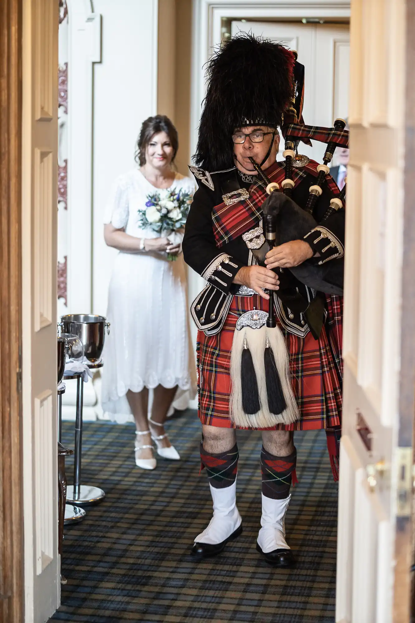 A man in traditional Scottish attire plays the bagpipes in the foreground. Behind him, a woman in a white dress holding a bouquet stands smiling in a doorway, suggesting a wedding or formal event in an indoor setting with tartan carpet.