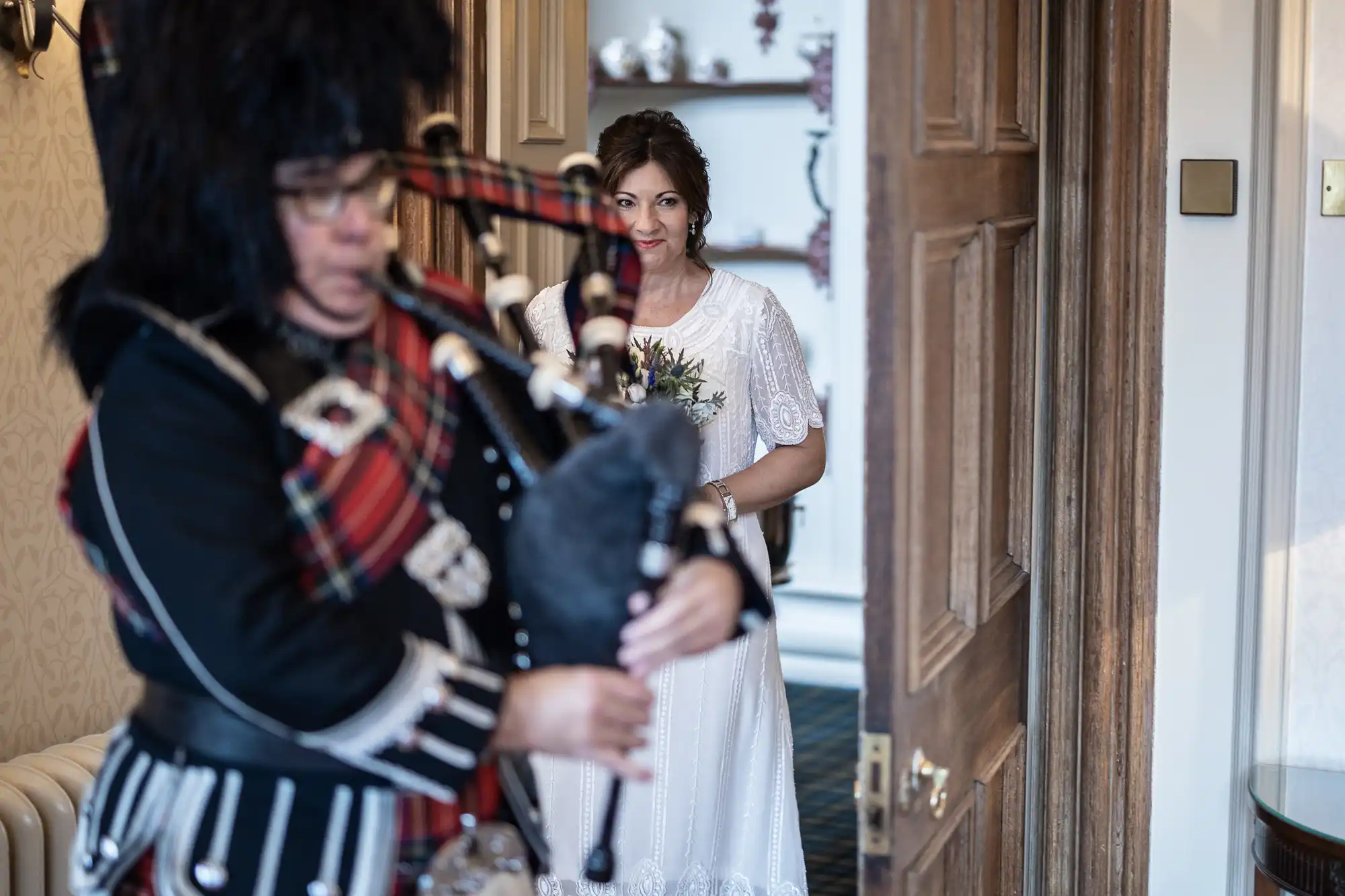 A bride in a white dress, holding a bouquet, stands smiling in a doorway. In the foreground, a person in traditional Scottish attire plays bagpipes, partially out of focus. The setting appears to be indoors, with wooden doors and shelves in the background.