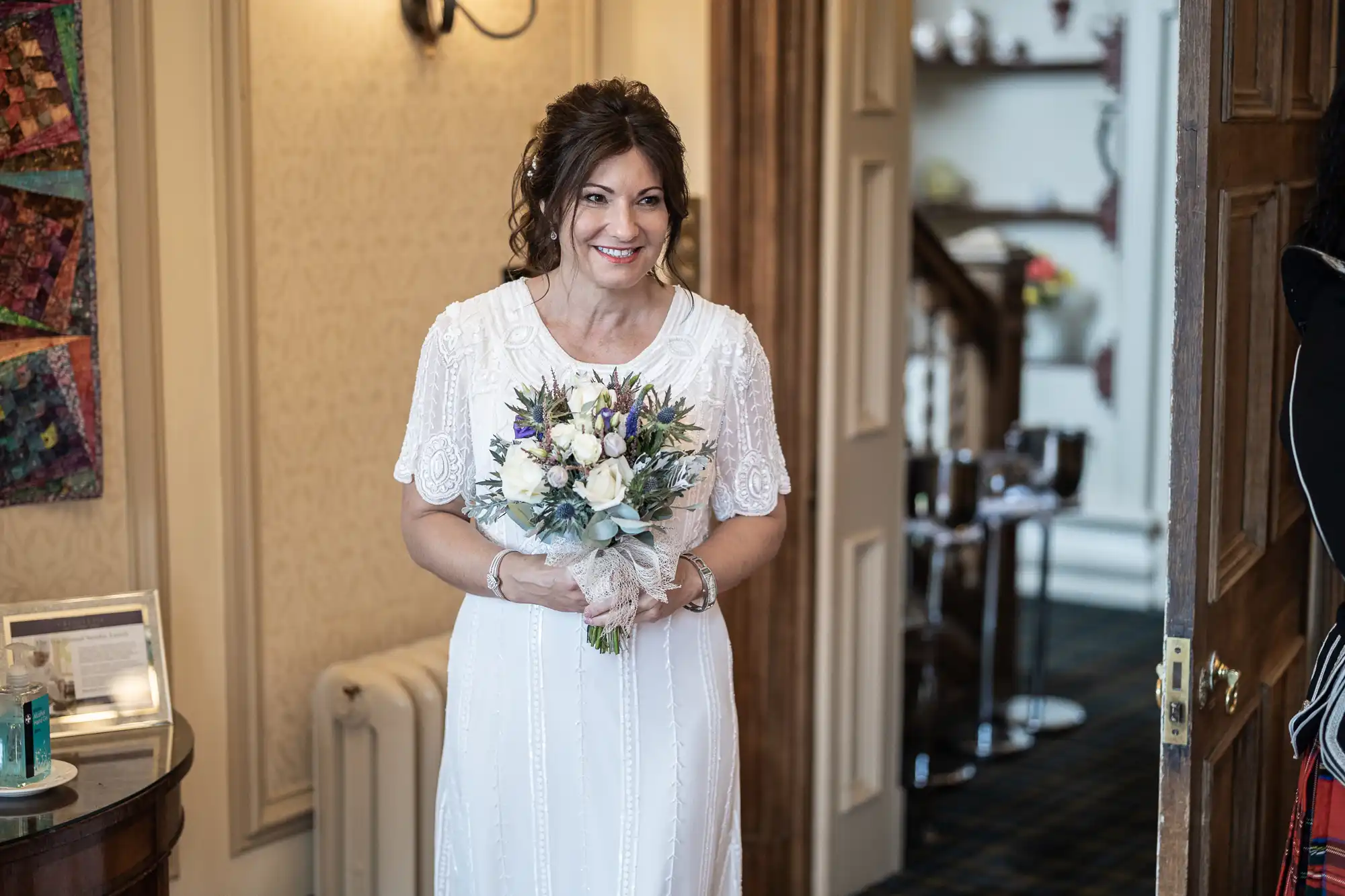 A smiling woman in a white, lace wedding dress holds a bouquet of white and purple flowers. She stands indoors near a doorway, with elegant decor, framed art, and a staircase visible in the background.