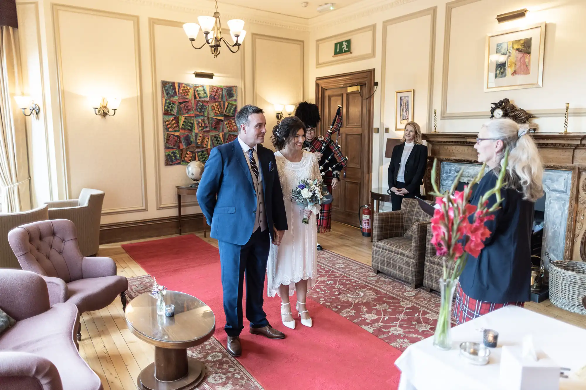A couple stands side by side, facing a woman officiant in a bright, elegant room with wooden floors, armchairs, and art on the walls. The woman wears a white dress and holds flowers; the man is in a suit. Three other people stand nearby, watching the ceremony.