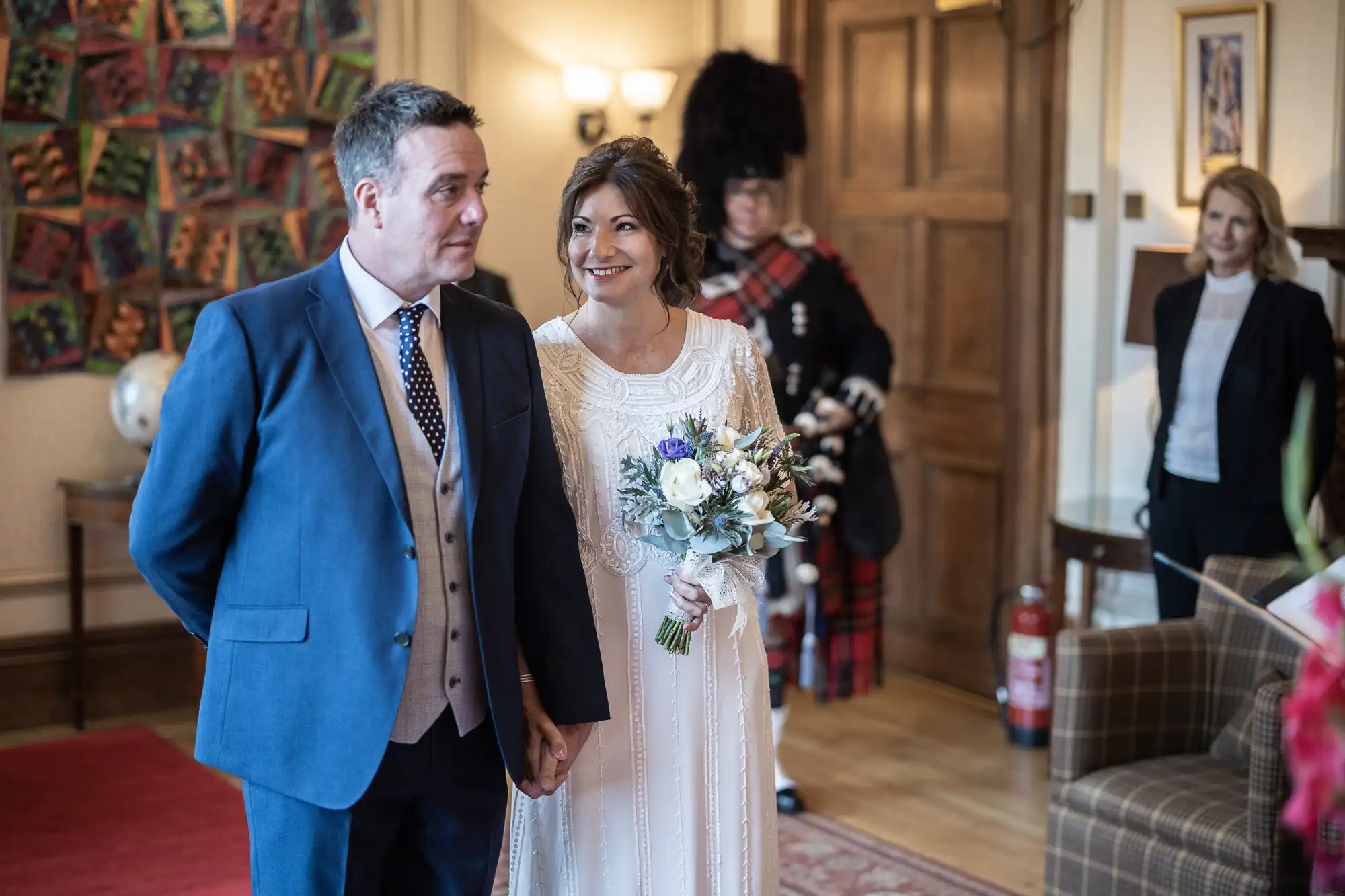 A bride in a white dress holding a bouquet stands, smiling at a man in a blue suit, holding his hand. Behind them is a person in traditional Scottish attire with a feathered hat, and a woman in black and white near an armchair in a warmly lit room.