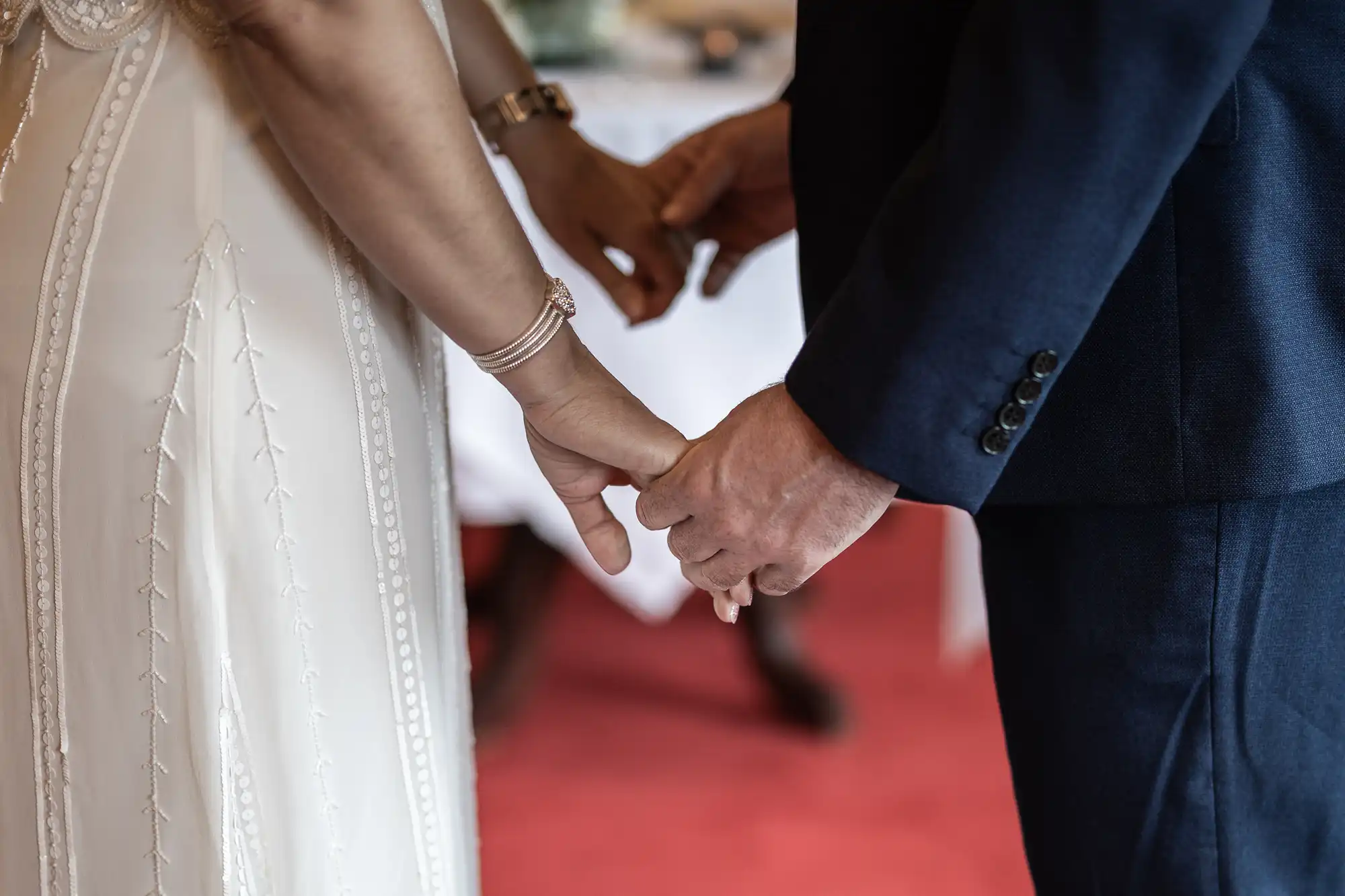A close-up of a couple holding hands. One person wears a white dress with decorative stitching and bracelets; the other wears a dark blue suit. The focus is on their joined hands, with a blurred background featuring a red carpet and white tablecloths.