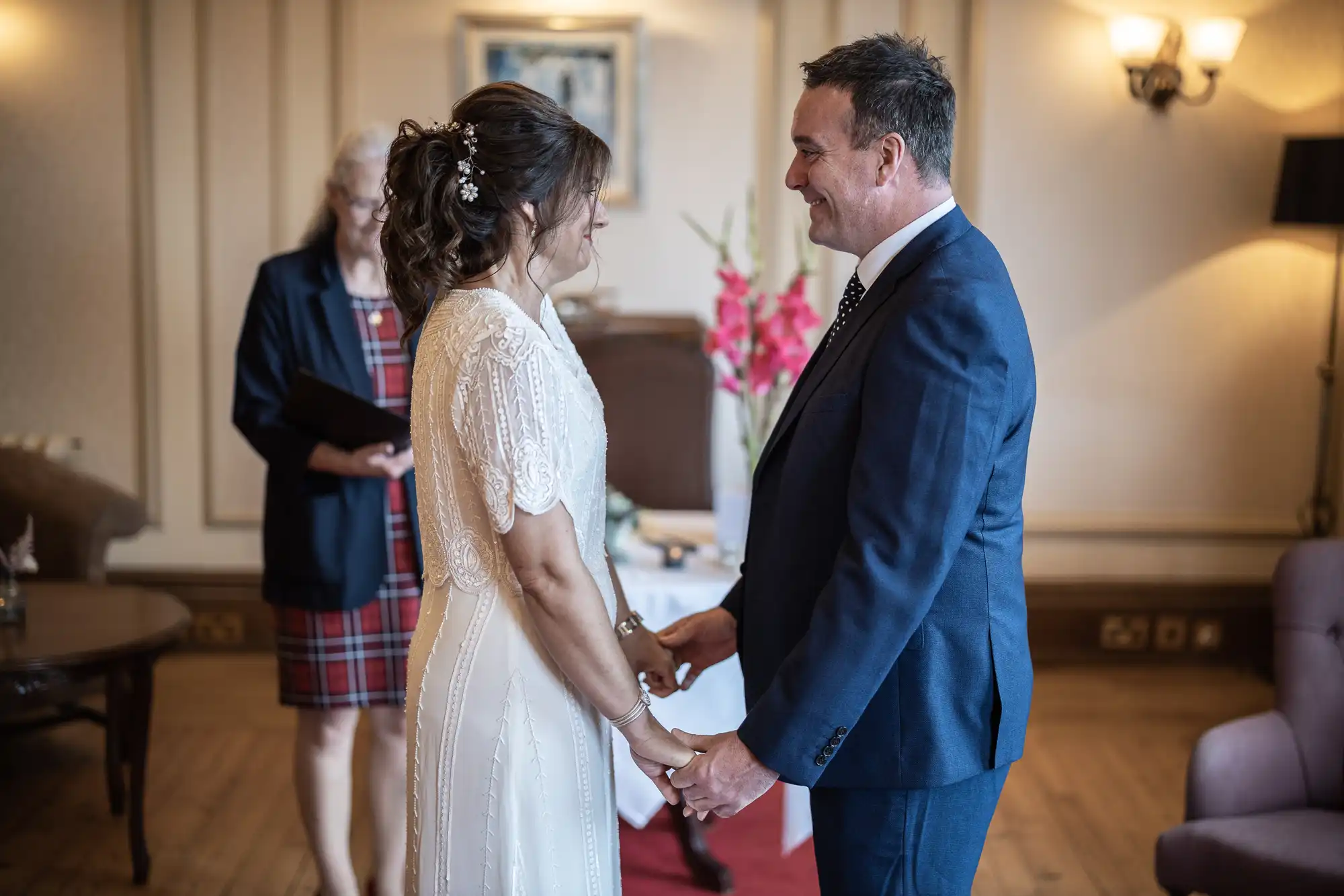 A bride and groom stand indoors, holding hands and smiling at each other during their wedding ceremony. The bride wears a white lace dress; the groom is in a navy suit. An officiant stands behind them in a checked dress and dark blazer. The room has elegant decor and flowers.