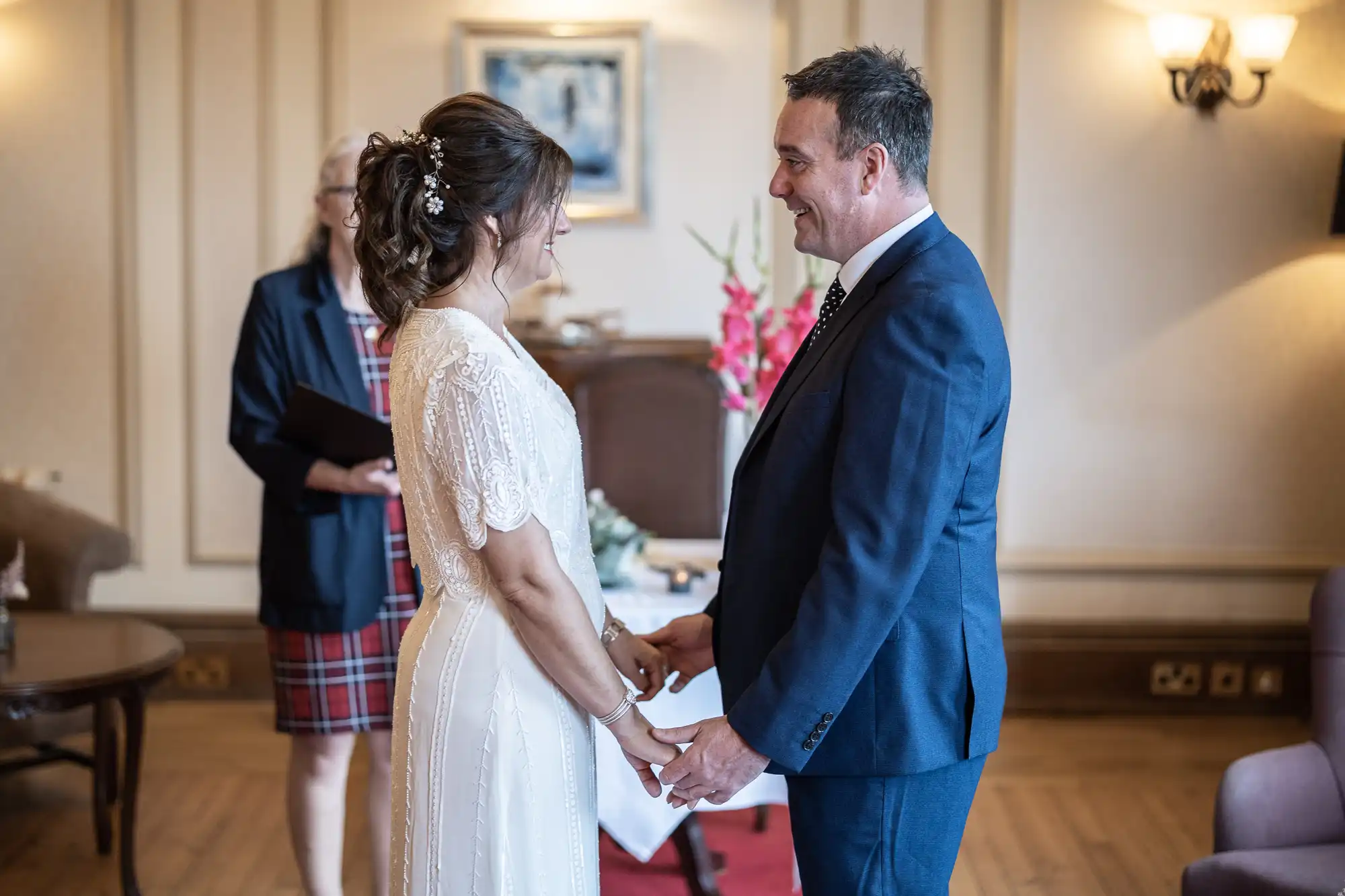 A bride in a white dress and a groom in a dark suit hold hands, smiling at each other during a wedding ceremony in an elegant room. A woman in a checked dress stands in the background, likely officiating. Flowers and lamps decorate the space.