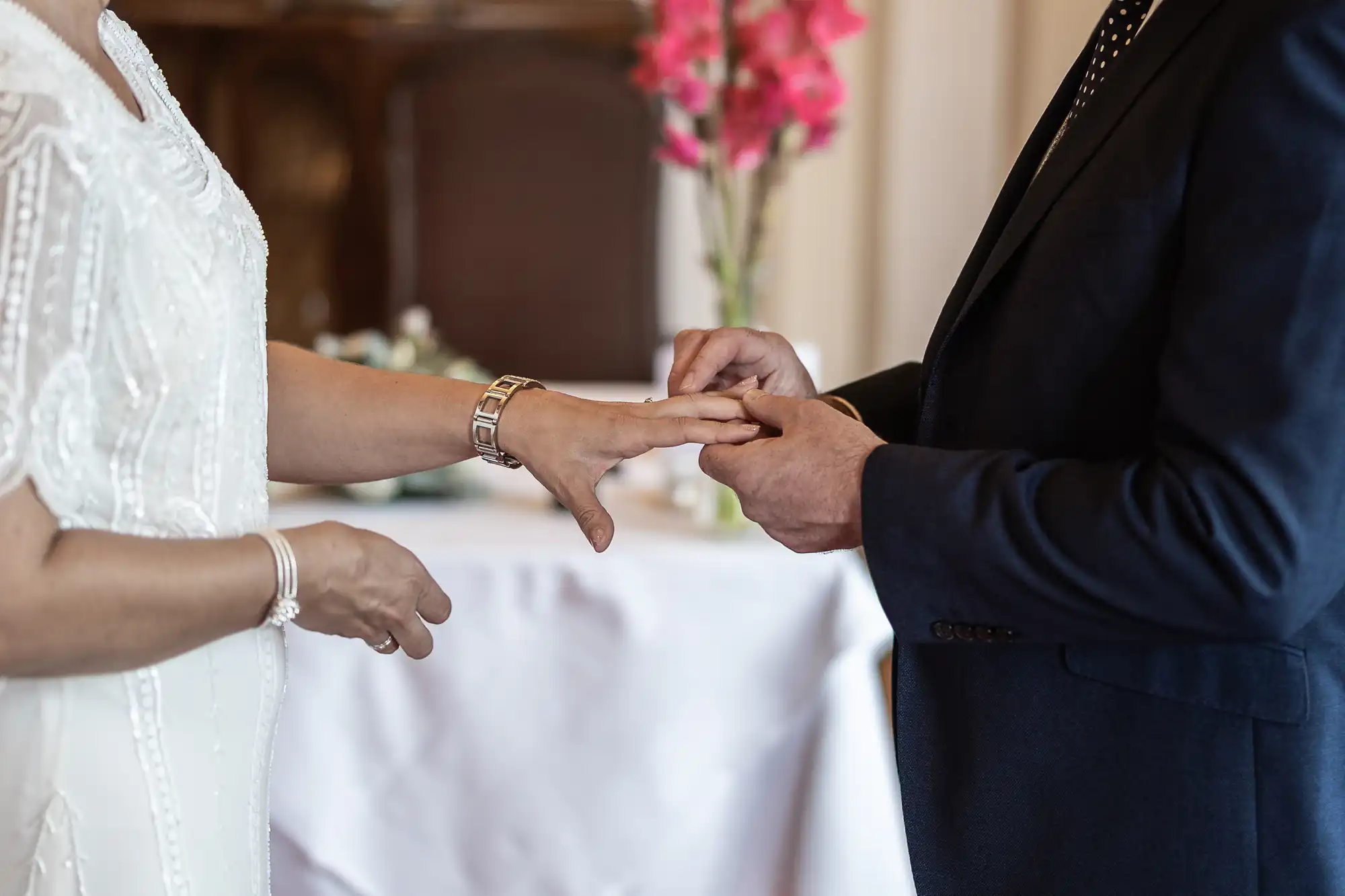 A person in a white dress and another in a dark suit exchange rings, with one slipping a ring onto the other's finger. Pink flowers and a white tablecloth are blurred in the background, suggesting a wedding or formal ceremony.