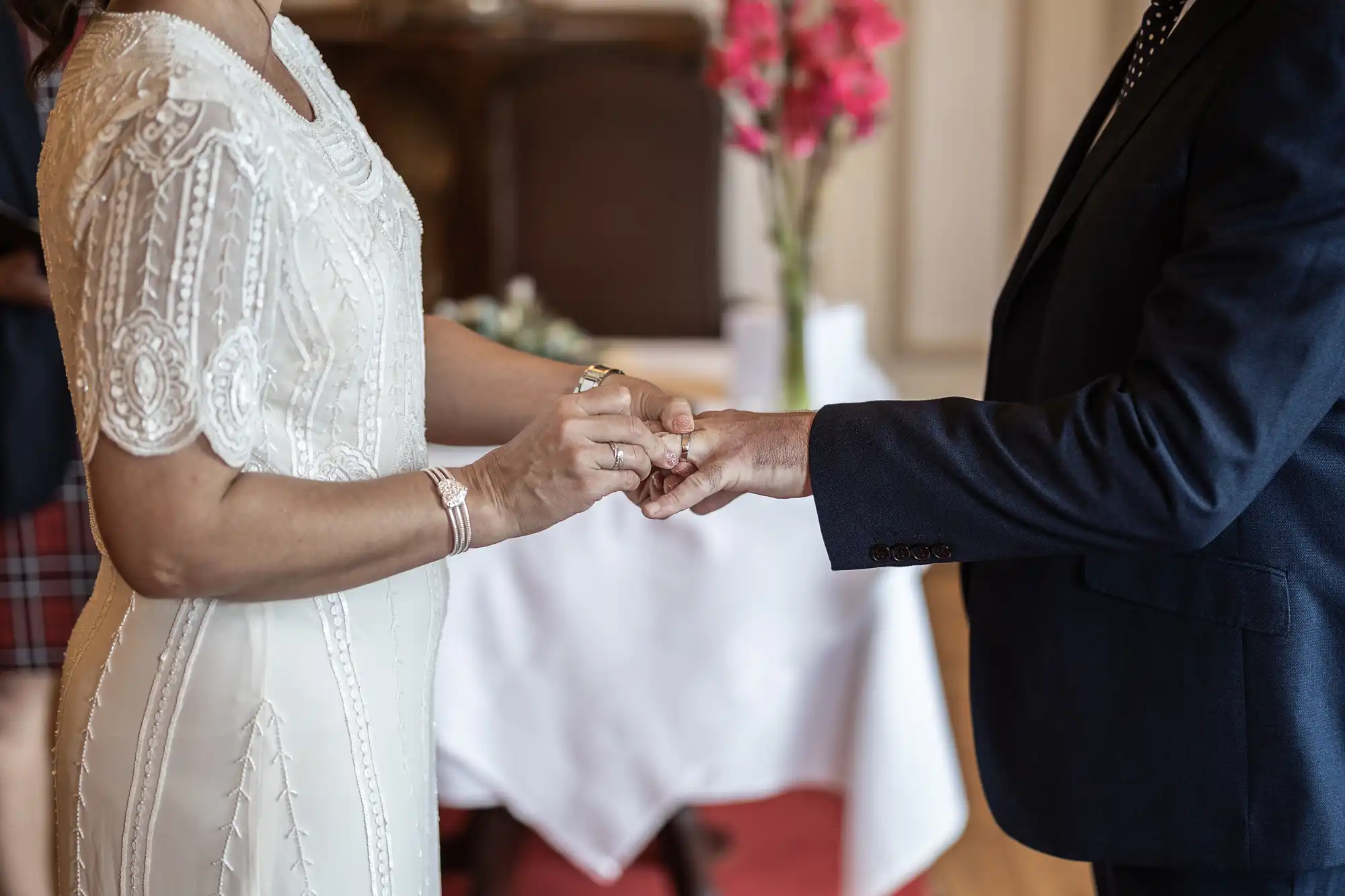 A couple stands facing each other, holding hands. The woman wears a white, embroidered dress, and the man is in a dark suit. A table with a vase of pink flowers is blurred in the background. Only their torsos and hands are visible.