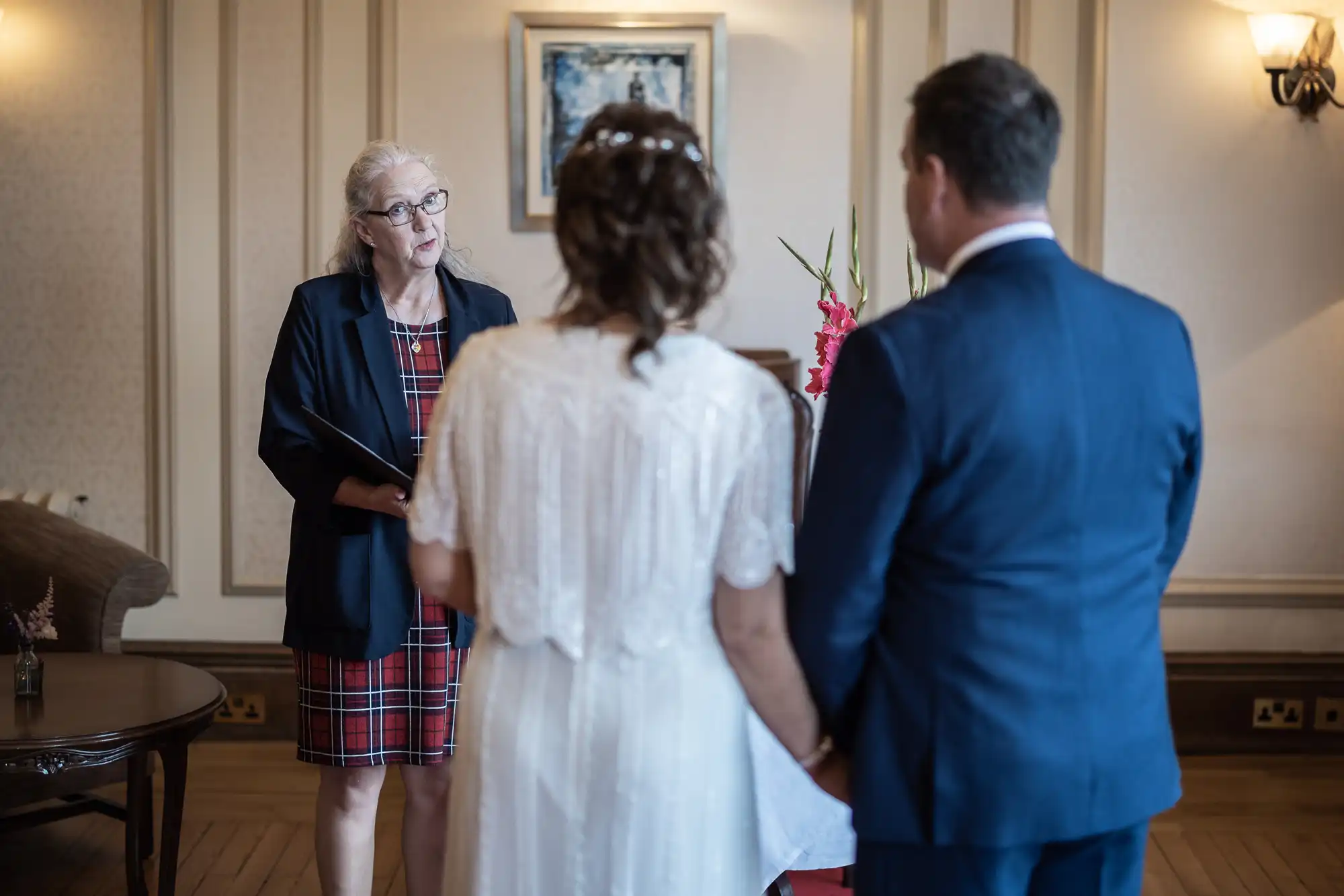 A woman officiant in glasses and a checked dress stands before a bride in a white dress and a groom in a blue suit, holding hands, during a wedding ceremony in an elegant, softly lit room with wood panelling and flowers.
