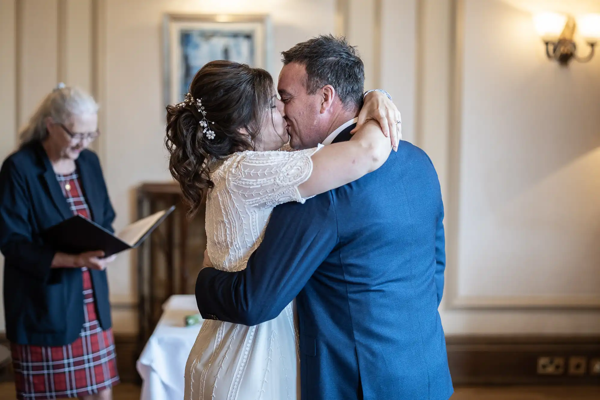 A bride in a white dress and a groom in a navy suit embrace and kiss indoors during their wedding ceremony, while an officiant holding a booklet stands nearby, reading. The room has cream-coloured walls, a framed picture, and a lit wall lamp.
