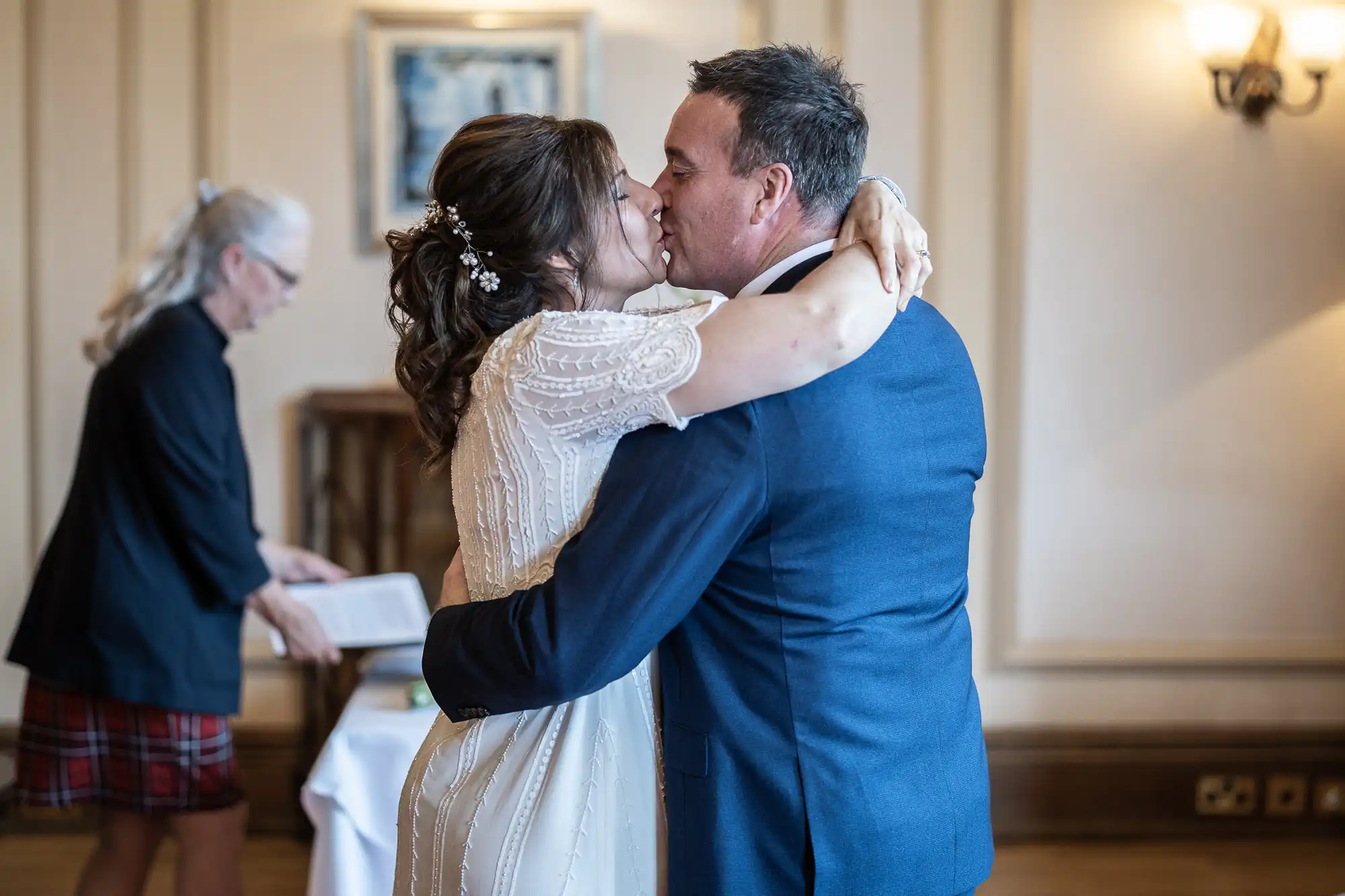 A bride in a white dress and a groom in a blue suit embrace and kiss indoors, celebrating their wedding. In the background, a woman with grey hair, glasses, and a checked skirt looks at a book or document, possibly officiating the ceremony.