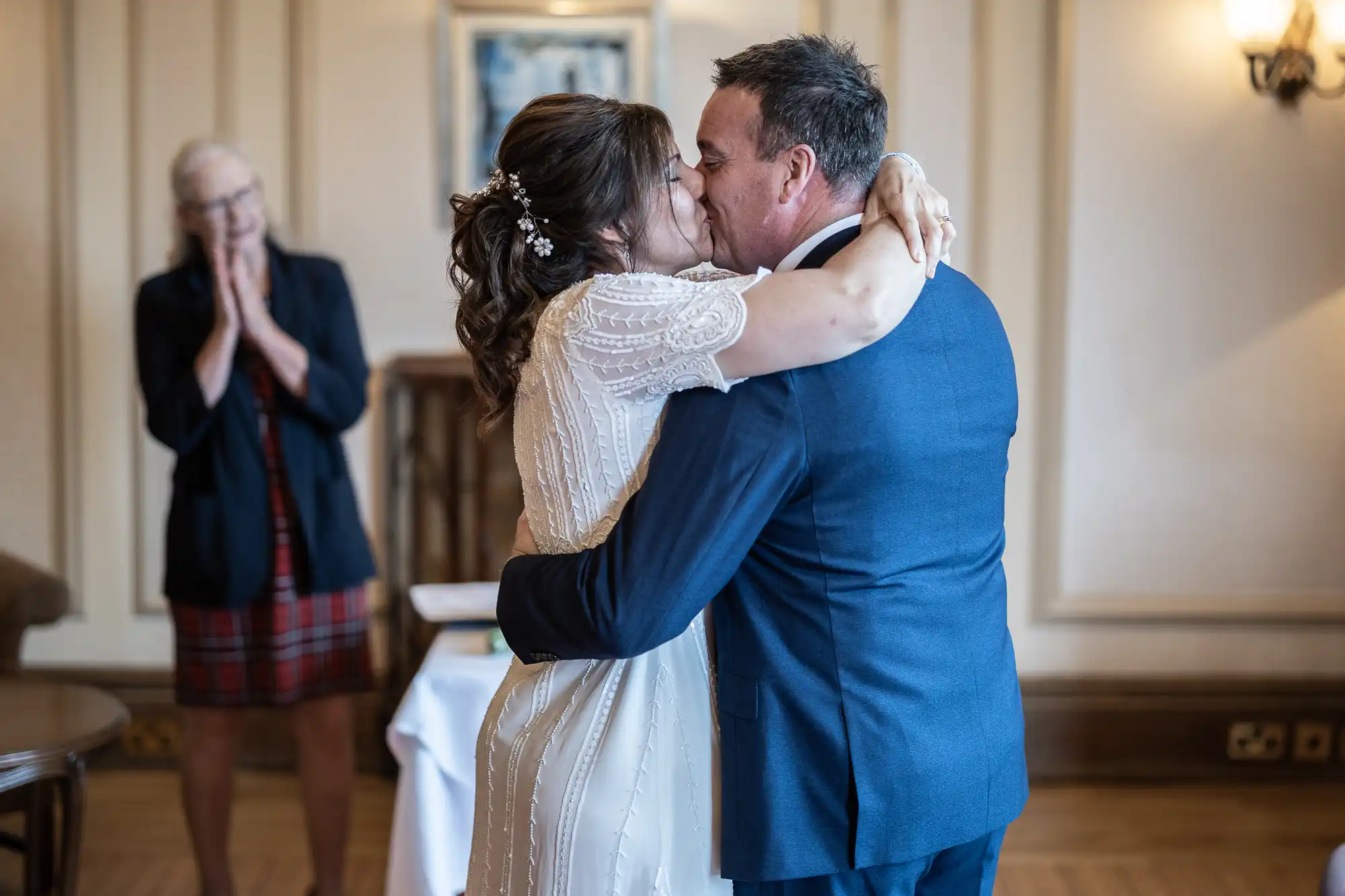 A bride in a white dress and a groom in a blue suit share a kiss while embracing indoors. A woman in a checked skirt and navy blazer stands in the background, smiling and clasping her hands in joy. The setting is warmly lit with cream-coloured walls.