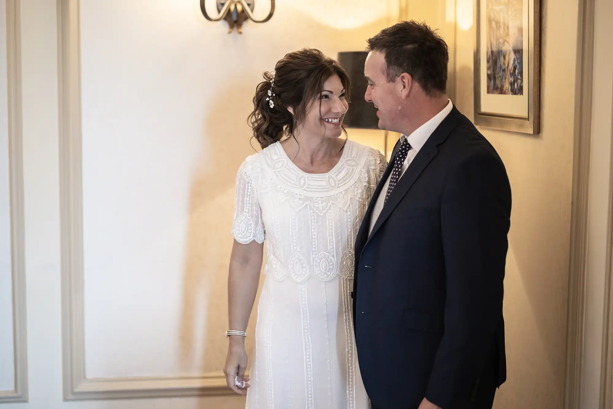A woman in a white, patterned dress and a man in a dark suit and spotty tie smile warmly at each other indoors; soft lighting and framed wall art create a cosy, elegant atmosphere.