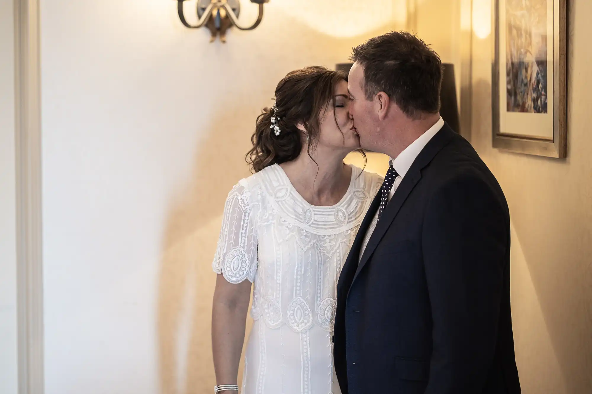 A bride in a white lace dress and a groom in a dark suit kiss indoors under a wall light, with a framed picture hanging on the cream-coloured wall beside them. The bride's hair is styled with small white flowers.