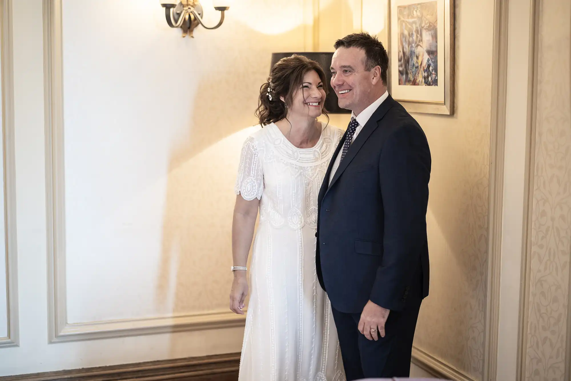 A woman in a white dress stands beside a man in a navy suit and tie. They are smiling and standing close together in a softly lit, elegant room with cream-coloured walls, a sconce, and a framed painting on the wall.