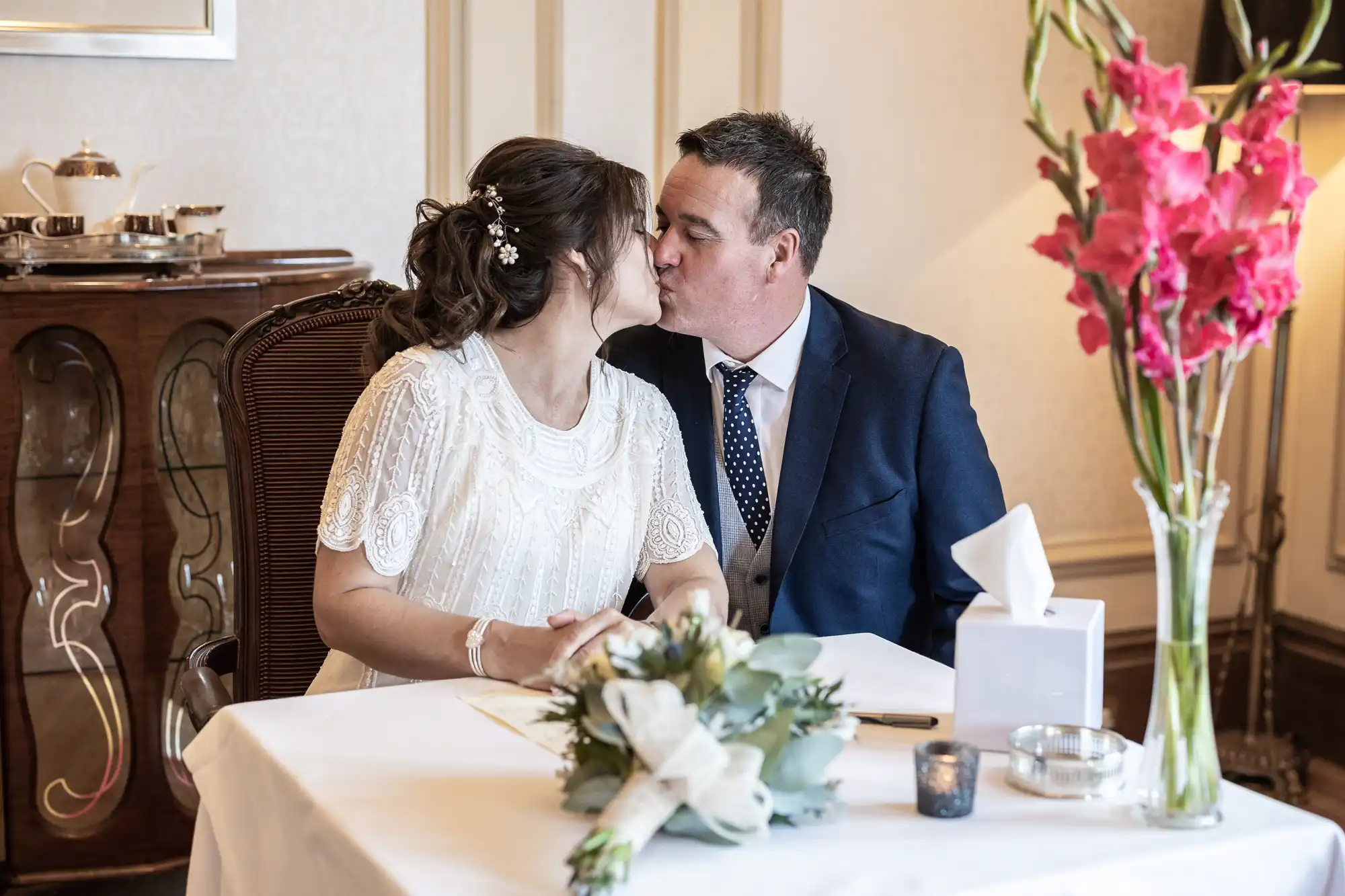 A bride and groom sit at a table, sharing a kiss. The bride wears a white dress with floral details; the groom wears a navy suit. A bouquet, pink flowers in a vase, a box of tissues, and papers are on the table. The setting is elegant and intimate.