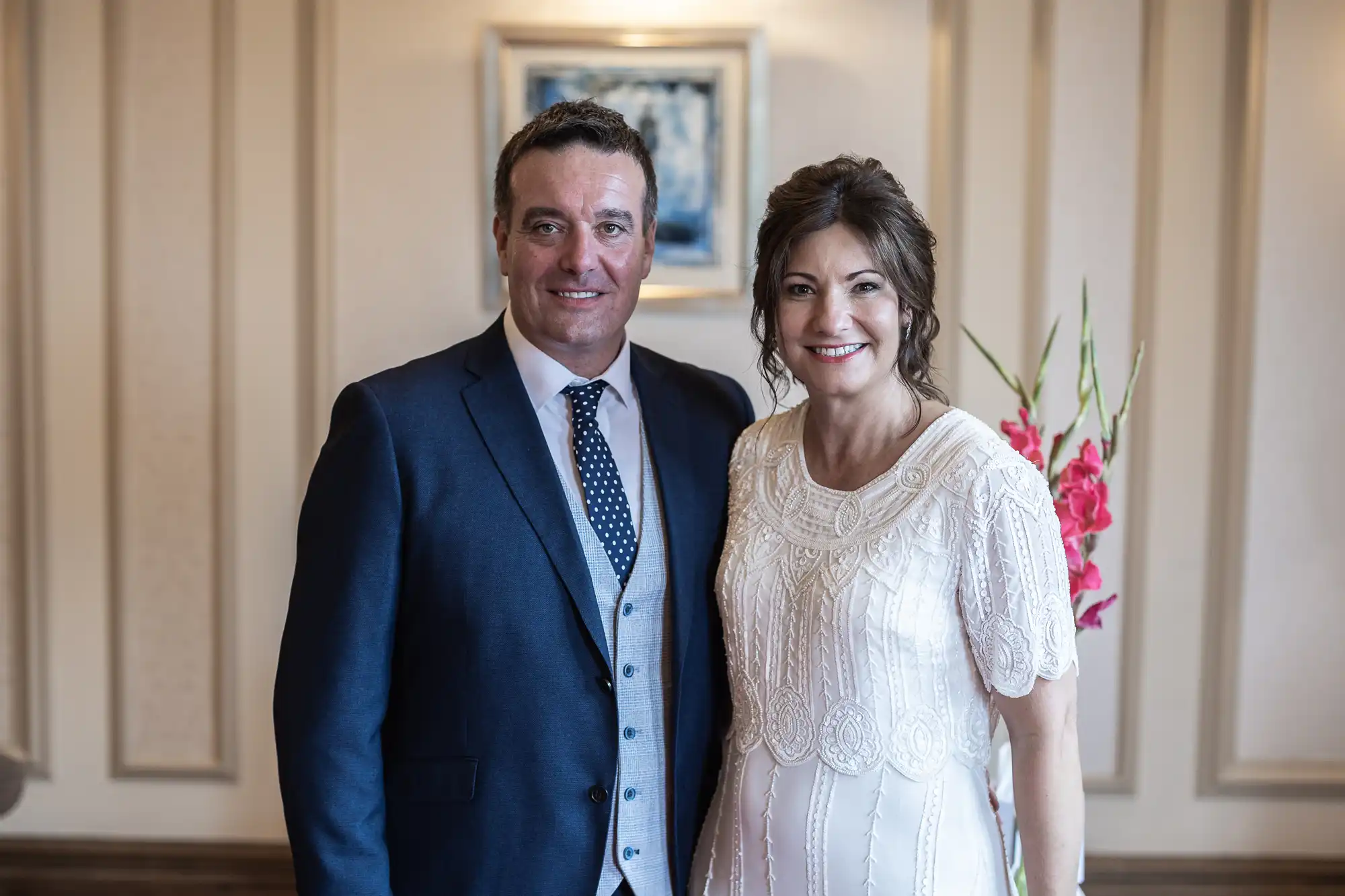 A man in a navy suit and polka-dot tie stands next to a woman in an elegant white dress. They are smiling and posing indoors. Behind them is beige panelling, a painting on the wall, and pink flowers on the right.