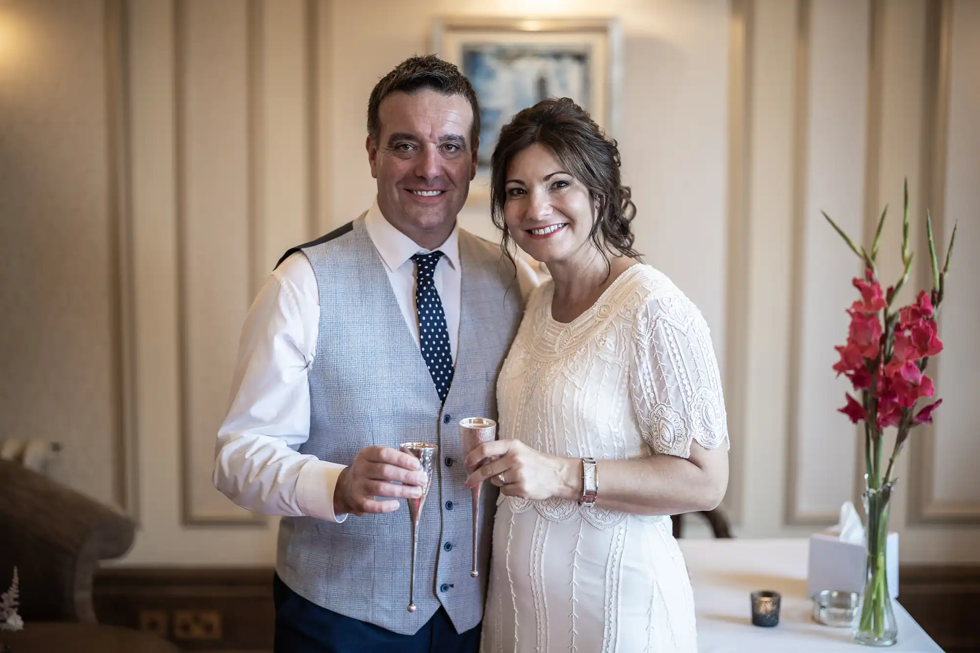 A smiling couple stands close together indoors, dressed in wedding attire. The man wears a light grey waistcoat, white shirt, and spotted tie. The woman wears a white, short-sleeved dress. Both hold small glasses. Red flowers and a candle are on the table beside them.