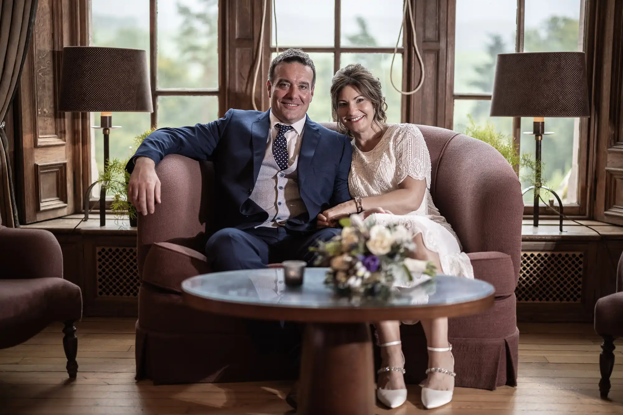 A smiling couple sit on a pink sofa in a wood-panelled room with large windows. The man wears a blue suit and the woman wears a white dress and heels. A bouquet and candle sit on a glass coffee table in front of them.