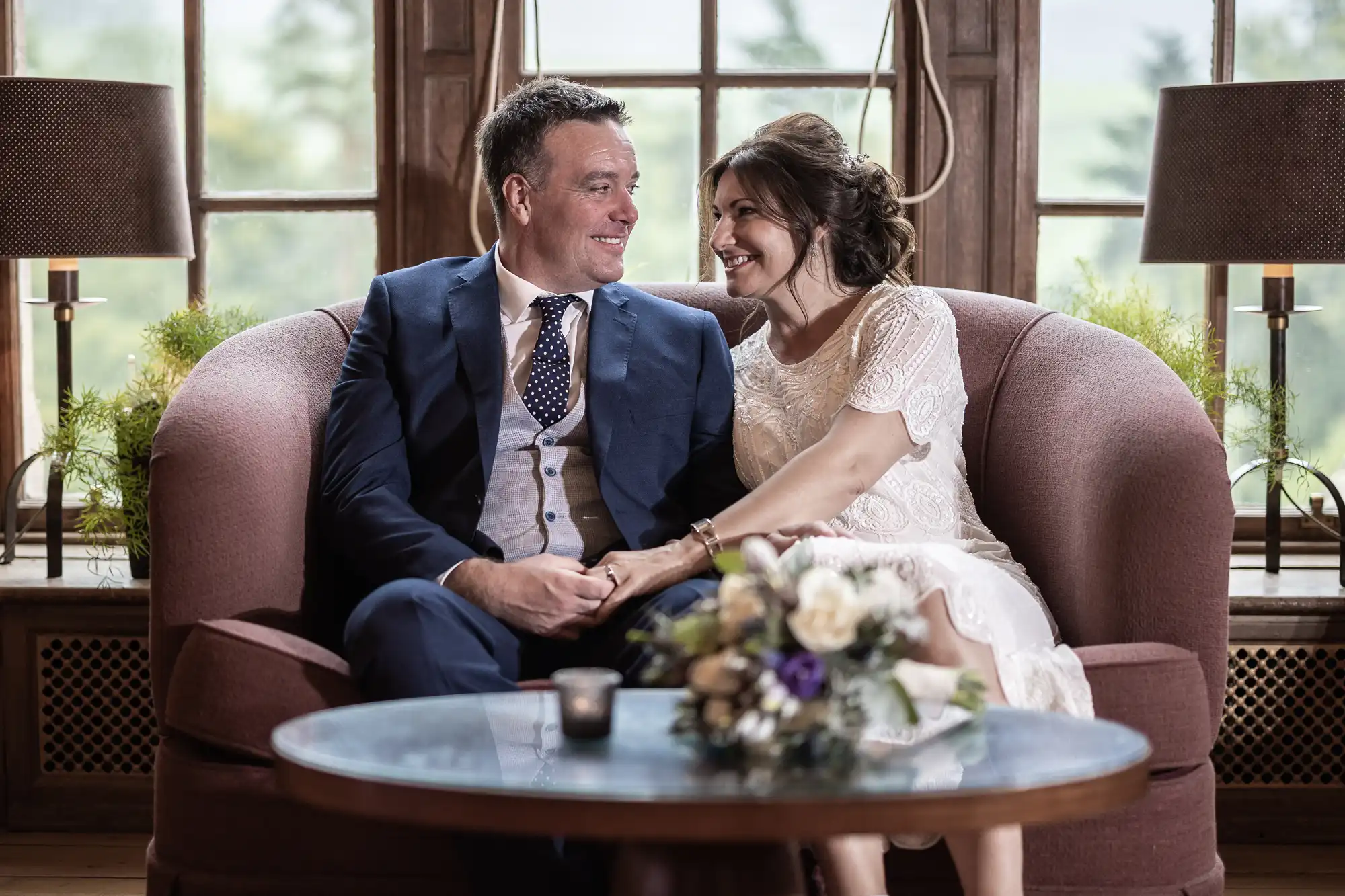A couple dressed in wedding attire sit close together on a mauve sofa, smiling at each other. Behind them are large windows with greenery outside. A glass table with a flower bouquet and candle is in the foreground. The mood is joyful and intimate.
