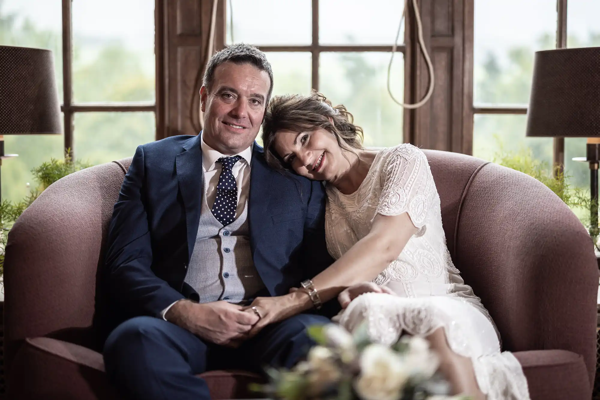 A man in a navy suit and a woman in a white lace dress sit closely on a mauve sofa. The woman leans her head affectionately on the man's shoulder, both smiling. Behind them is a window with natural light, and a bouquet is blurred in the foreground.