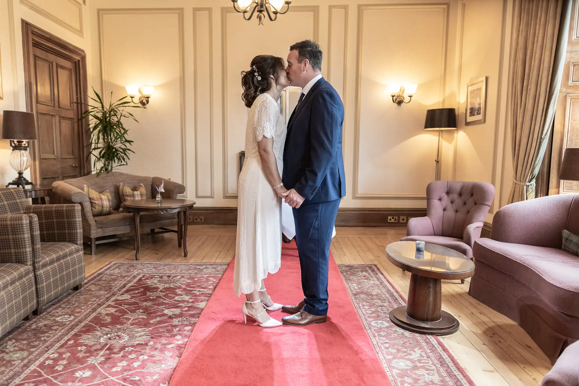 A bride in a white dress and groom in a blue suit stand face-to-face, holding hands and sharing a kiss on a red carpet in an elegant, warmly lit room with classic furniture and wooden floors.