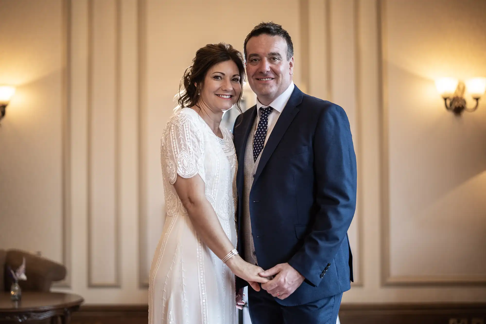 A smiling couple stands indoors, holding hands. The woman wears a white, short-sleeved, beaded dress; the man wears a navy suit with a blue polka-dot tie and light waistcoat. They stand close together in a softly lit, elegant room with cream-coloured walls.