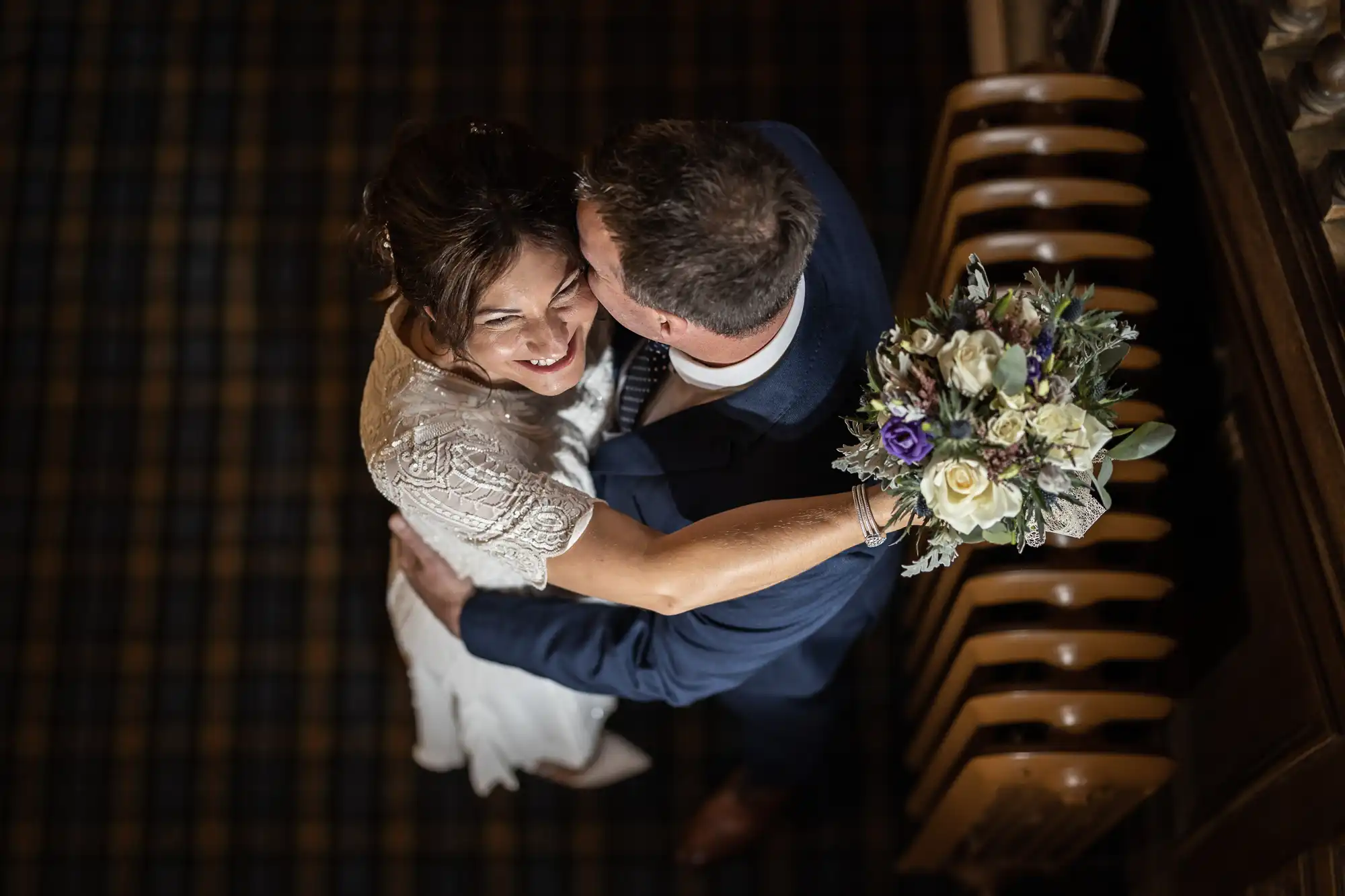 A bride in a white dress and groom in a dark suit embrace and smile, seen from above. The bride holds a bouquet of cream, purple, and green flowers. They stand close together on a dark, chequered floor next to stacked wooden chairs.