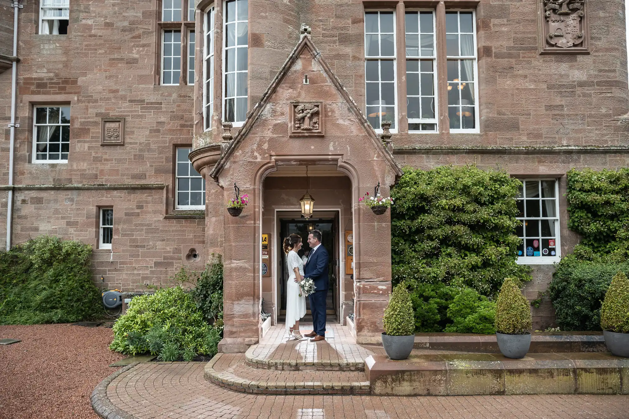 A bride and groom stand under the stone archway of a historic brick building, holding hands and smiling at each other. The entrance is adorned with hanging flower baskets, tall windows, green shrubs, and large potted plants on either side.