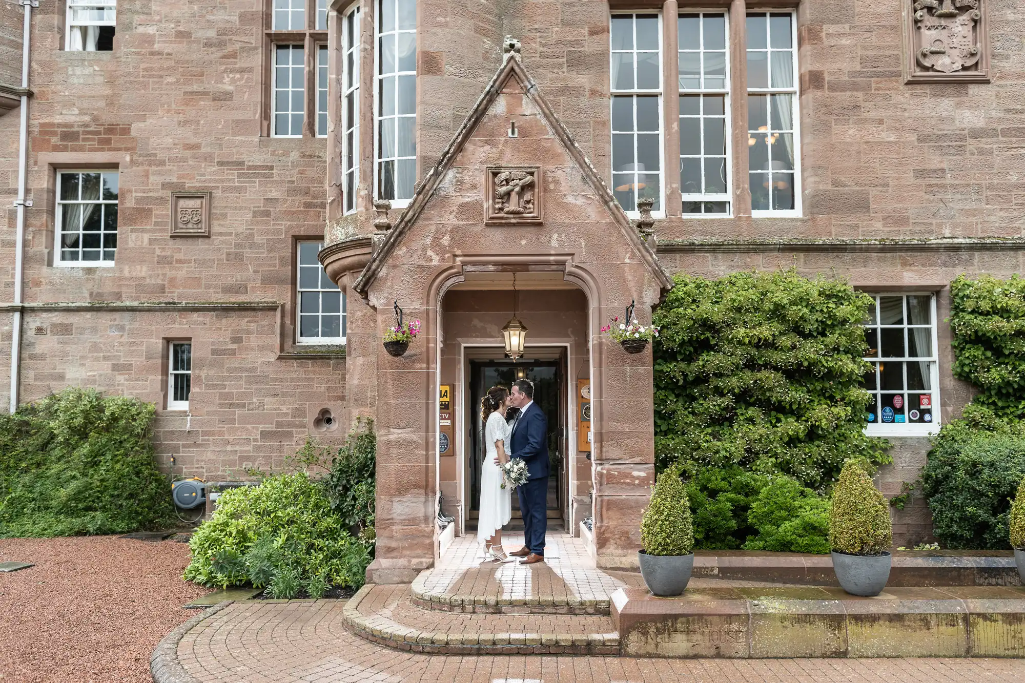 A bride and groom stand facing each other under the stone entrance of a historic building with large windows, greenery, and potted plants. They hold hands and smile, dressed in wedding attire. The scene is bright and celebratory.