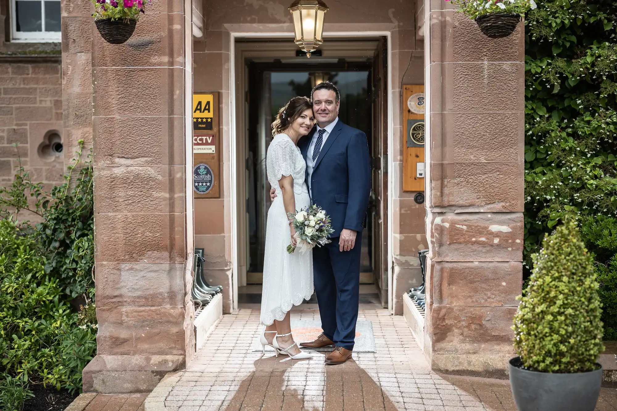 A bride in a white dress and a groom in a blue suit stand closely together, smiling, in a stone archway. The bride holds a bouquet and wears white heels. Hanging plants and green bushes frame the entrance behind them.