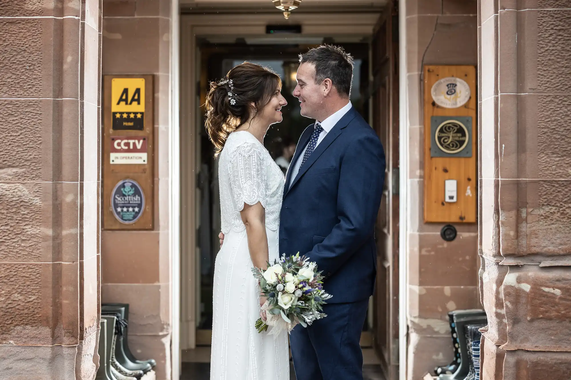 A bride in a white dress holding a bouquet and a groom in a navy suit smile and gaze at each other, standing in a stone doorway decorated with plaques. They are outdoors, surrounded by brown stone walls and old-fashioned benches.