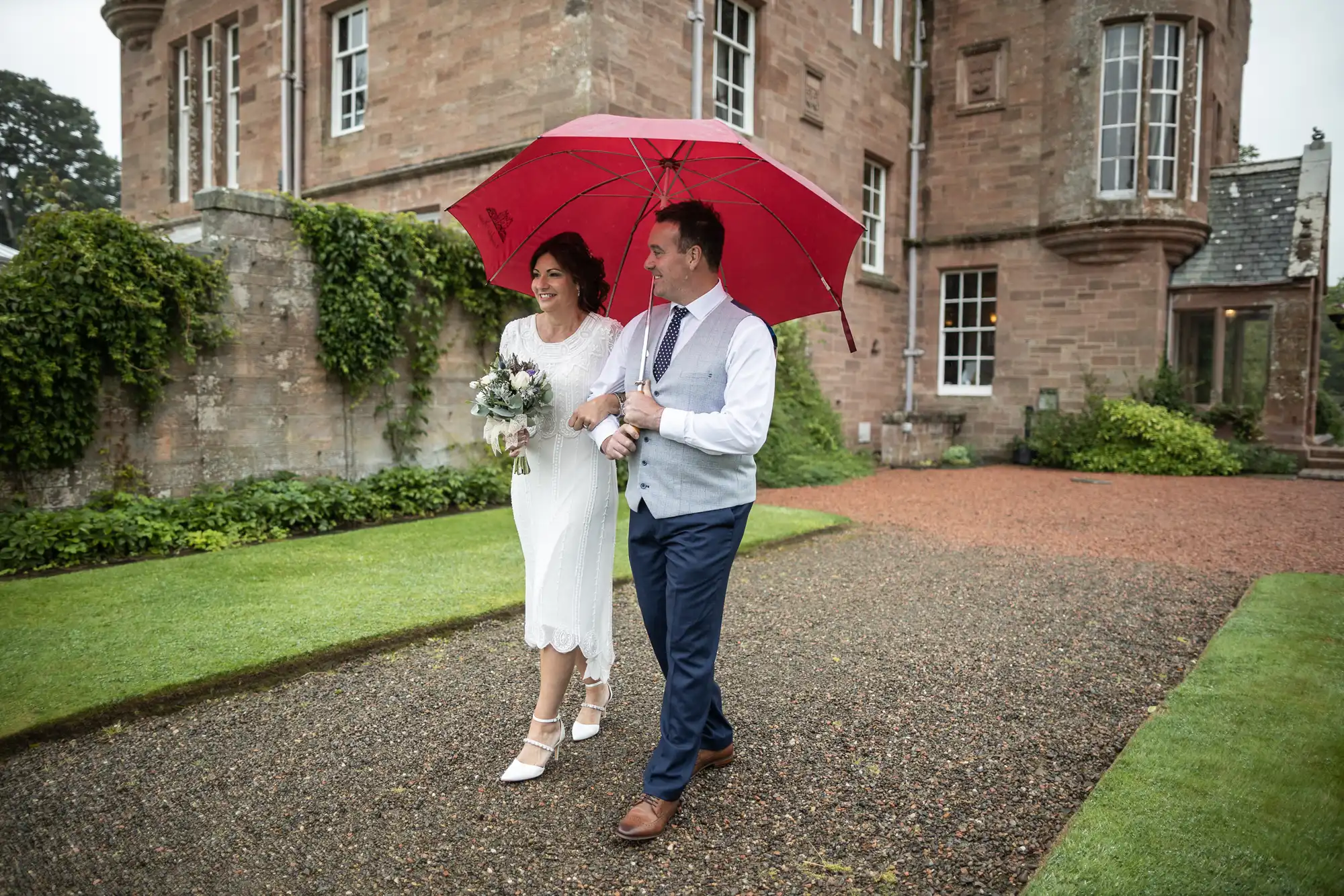 A bride in a white dress and groom in a waistcoat and tie walk arm-in-arm on a gravel path outside a stone building, smiling and sharing a large red umbrella. The bride holds a bouquet, and the grass and ivy-covered walls appear lush and green.