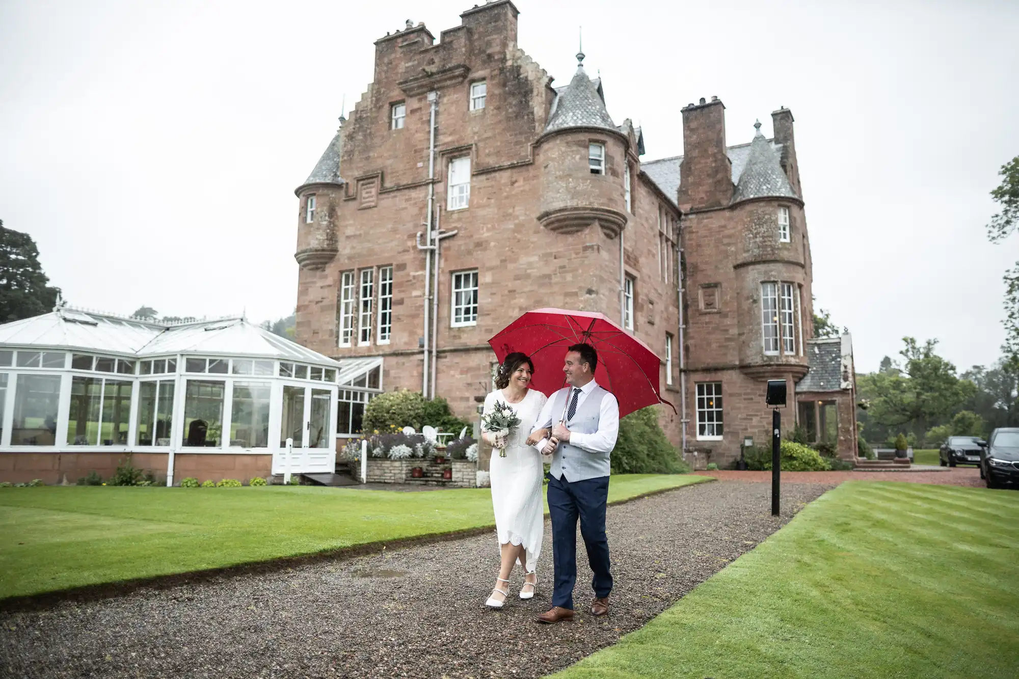 A bride and groom walk arm-in-arm on a gravel path outside a large, historic stone castle. The groom holds a red umbrella over them. The bride wears a white dress and holds a bouquet. The grass is green, and the sky is overcast, suggesting rain.