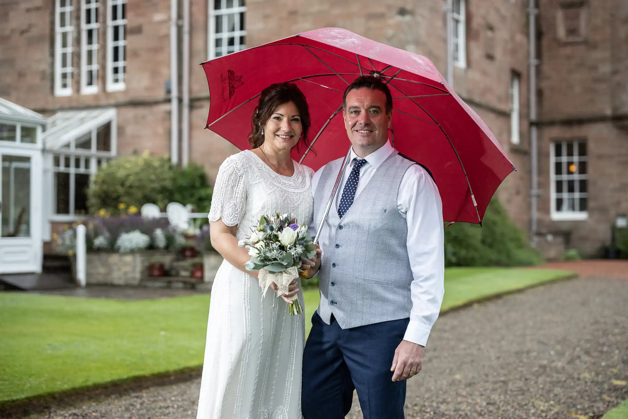 A smiling couple pose outdoors on a wedding day. The woman wears a white dress and holds a bouquet; the man wears a grey waistcoat, white shirt, and navy tie. They both stand under a large red umbrella in front of a stone building, with greenery in the background.