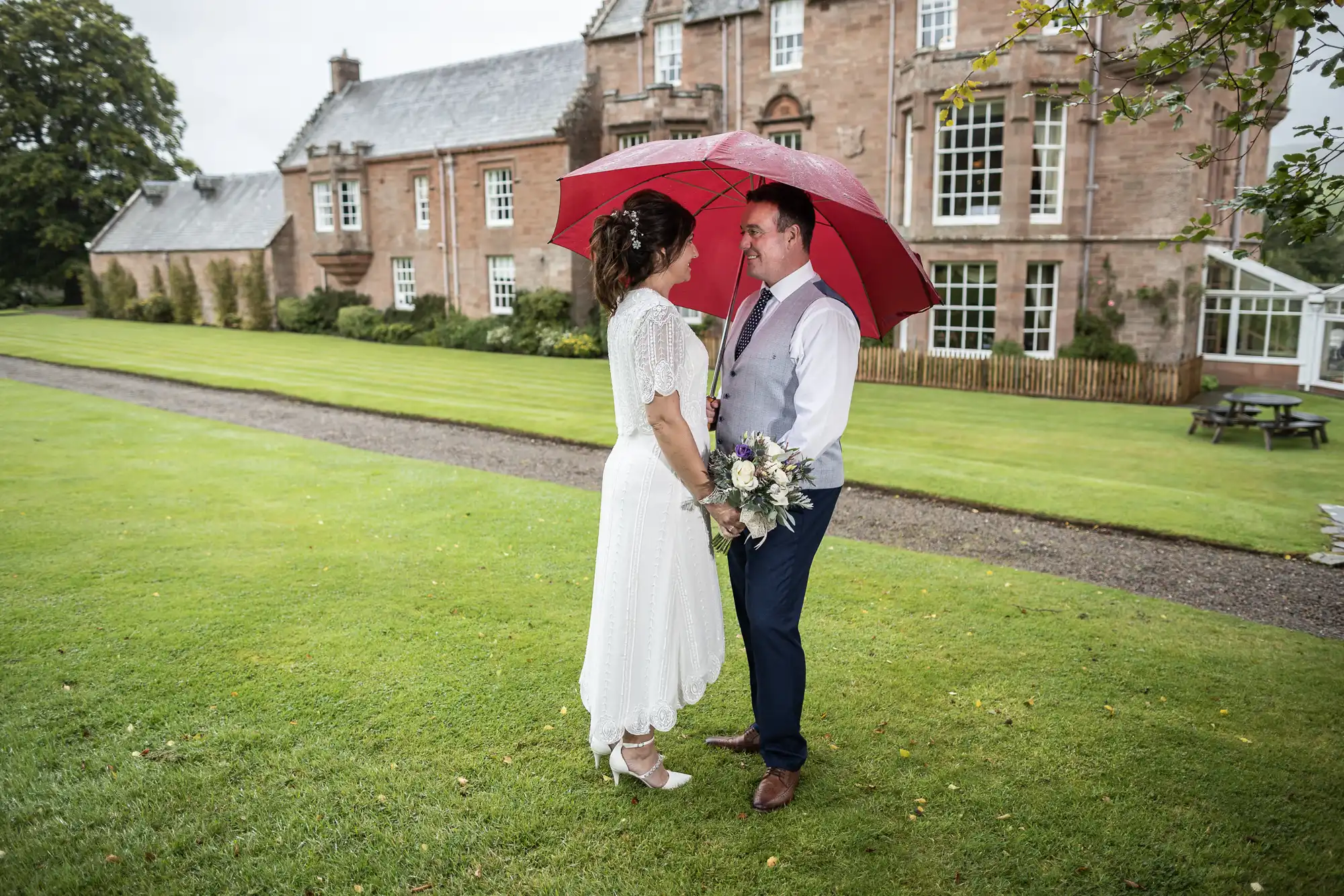A bride and groom stand facing each other under a red umbrella on a grassy lawn in front of a large stone building. The bride wears a white dress and holds a bouquet; the groom wears dark trousers and a waistcoat. It is overcast and the ground appears wet.