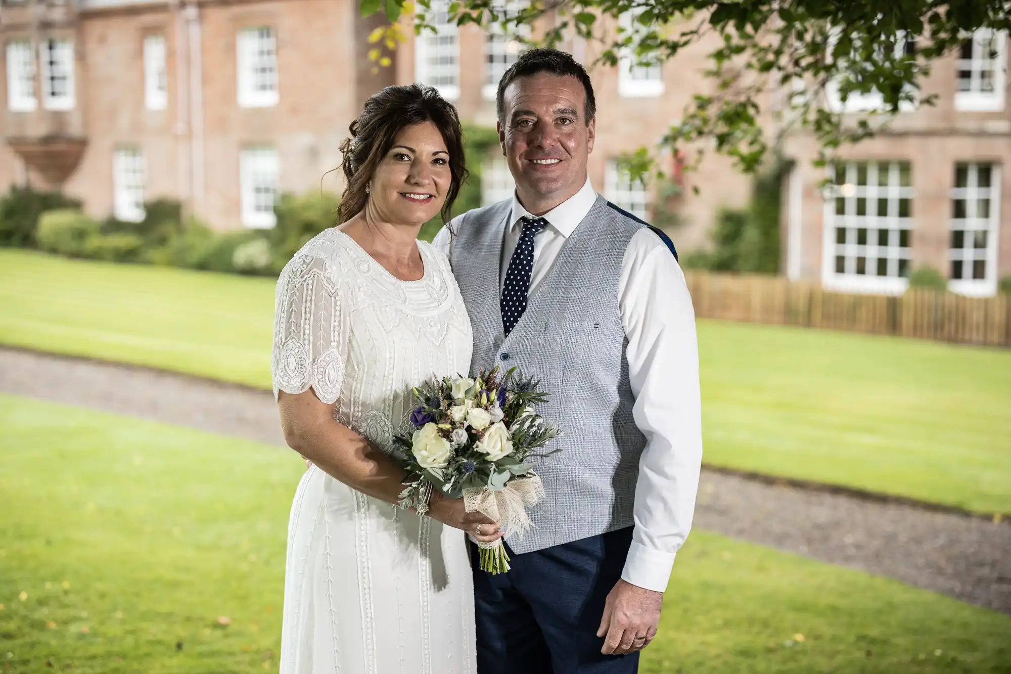 A smiling couple pose outdoors; the woman wears a white dress and holds a bouquet of flowers, whilst the man wears a light grey waistcoat, white shirt, and navy tie. They stand on green grass with a stately building and trees blurred in the background.