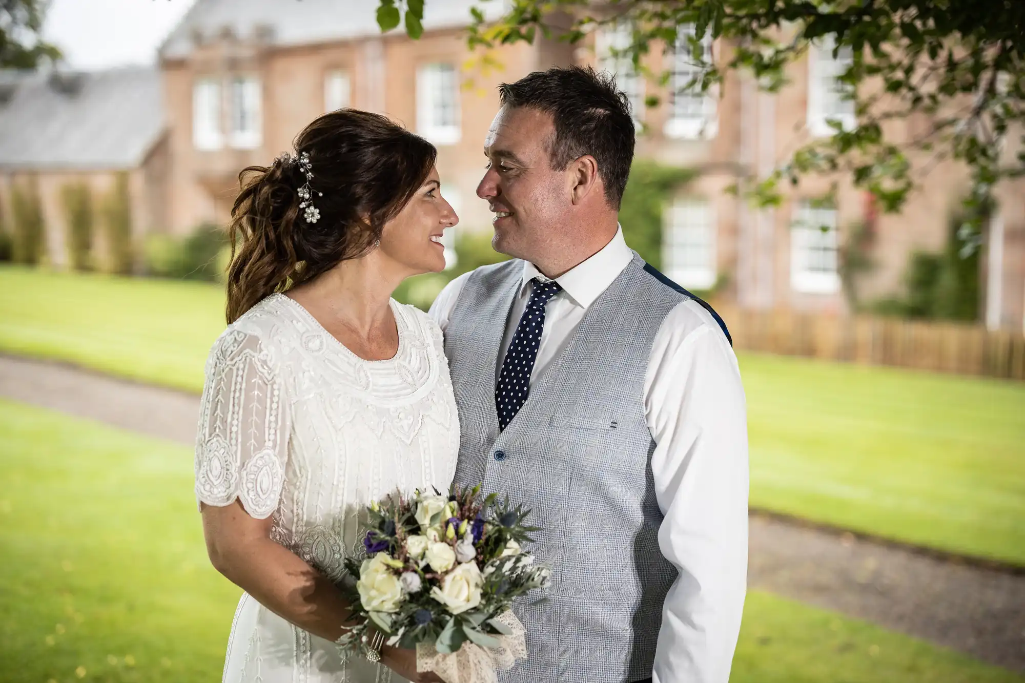 A bride in a white dress holding a bouquet and a groom in a waistcoat and tie stand outdoors, smiling at each other. They are in front of a brick building surrounded by green grass and trees, with soft daylight illuminating the scene.