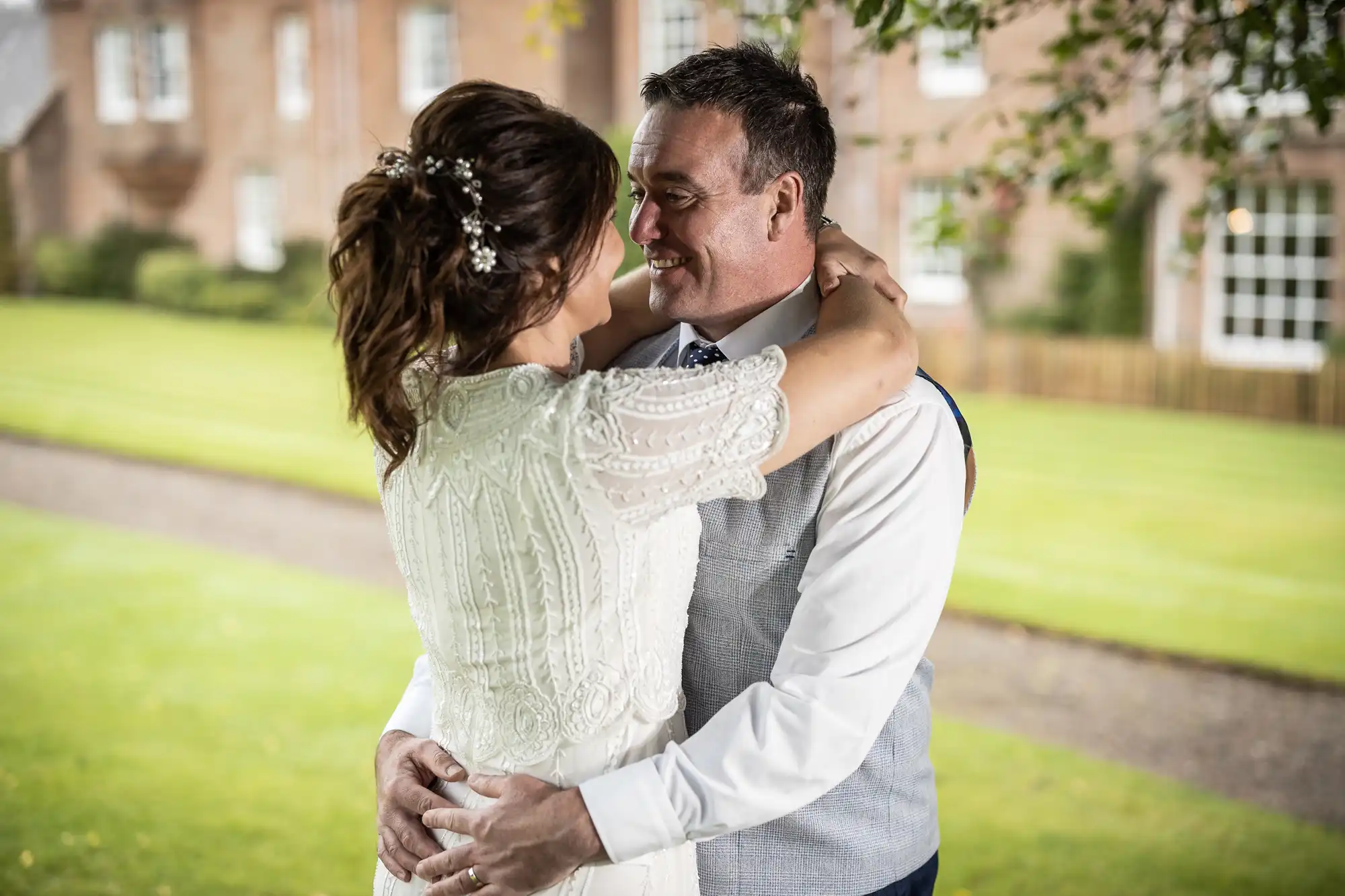 A bride and groom embrace outdoors, smiling at each other. The bride wears a white dress with beaded details and a floral hair accessory. The groom wears a white shirt, tie, and light grey waistcoat. Lush green grass and a brick building are in the background.