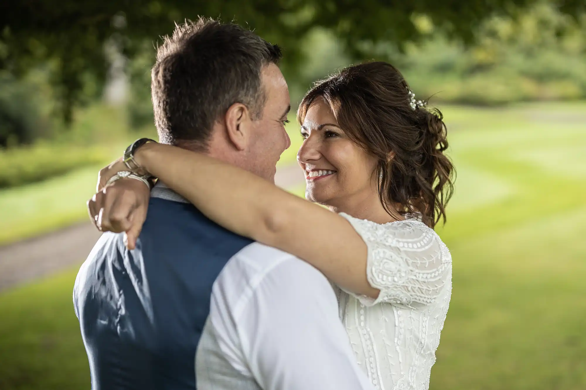 A bride and groom embrace outdoors, smiling at each other under a tree. The bride wears a white dress with lace details, her arms around the groom's neck. The groom, in a white shirt and blue waistcoat, faces her. A green lawn and pathway appear in the background.