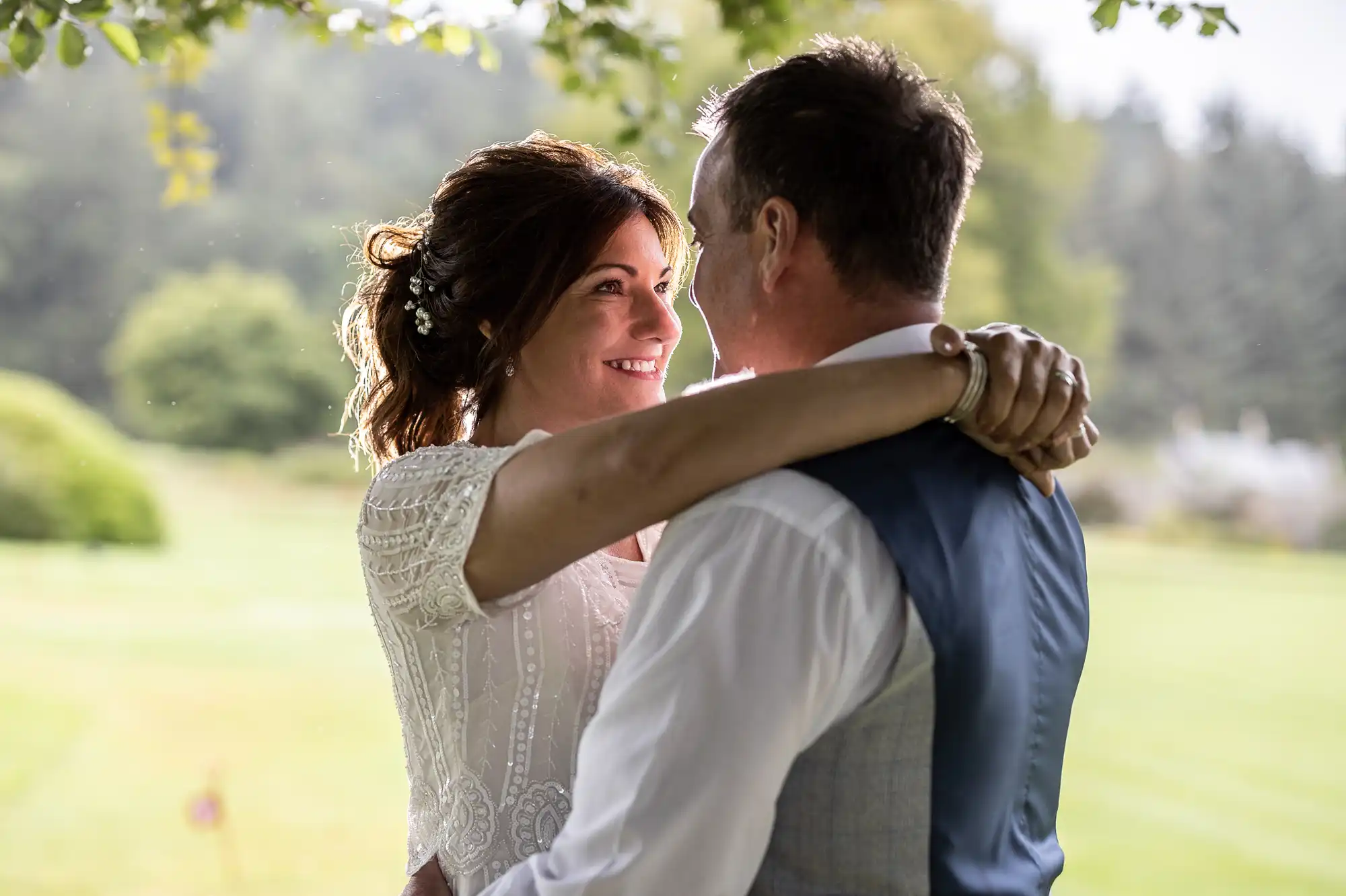 A bride and groom embrace outdoors, smiling and gazing into each other’s eyes. The bride wears a white dress with lace sleeves, and the groom wears a white shirt and grey waistcoat. Green trees and grass create a bright, blurred background.