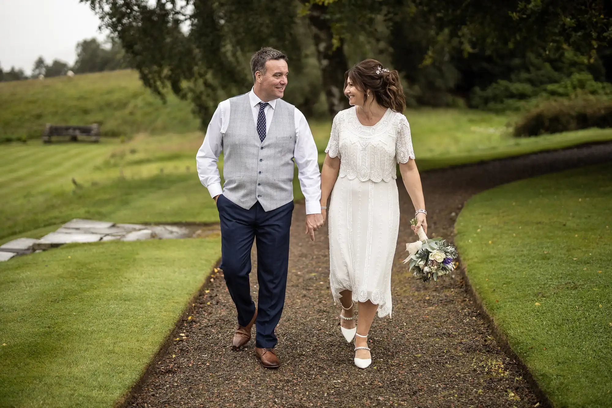 A smiling couple walk hand in hand along a garden path. The woman wears a white, knee-length dress and holds a bouquet, while the man wears blue trousers, a grey waistcoat, and a tie. Both look at each other, surrounded by green grass and trees.