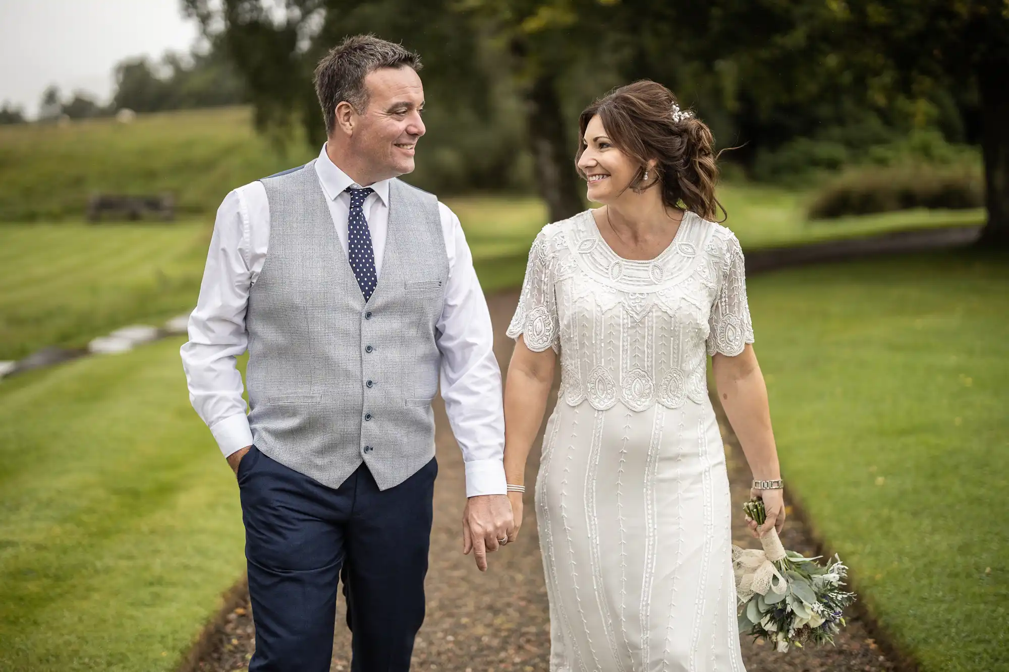 A couple walks hand-in-hand outdoors on a path. The man wears a light grey waistcoat, white shirt, navy trousers, and tie. The woman wears a white, beaded dress and holds a bouquet. Both smile warmly at each other, surrounded by lush green grass and trees.