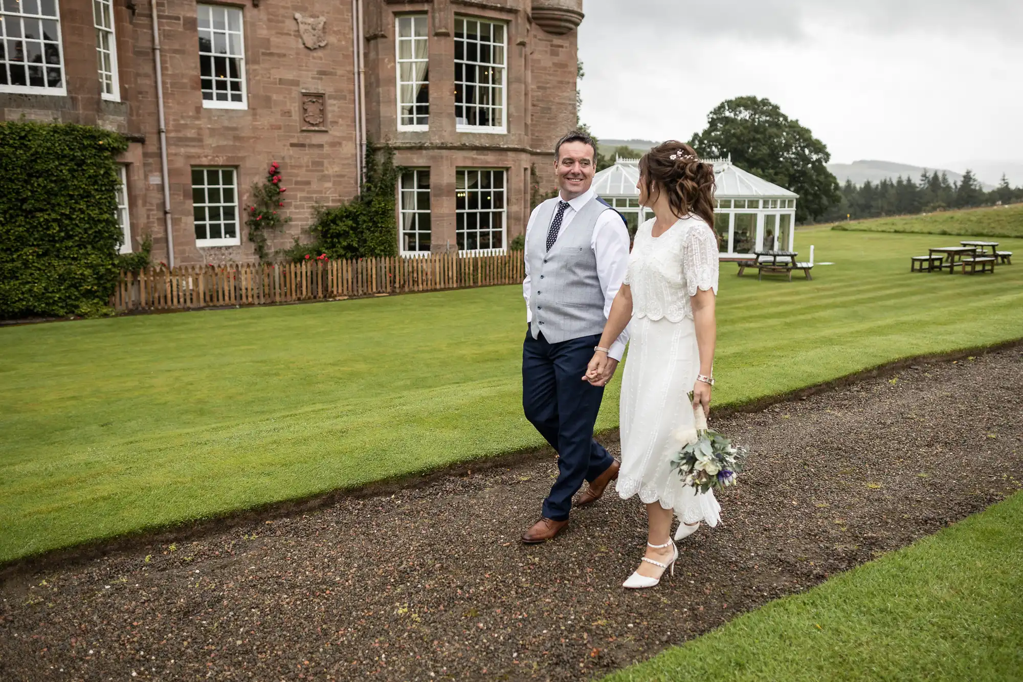 A couple walk hand-in-hand on a gravel path beside a large historic building and manicured lawn. The woman wears a white dress, holding a bouquet, and the man wears a waistcoat and tie. Both are smiling and looking at each other, with garden benches in the background.