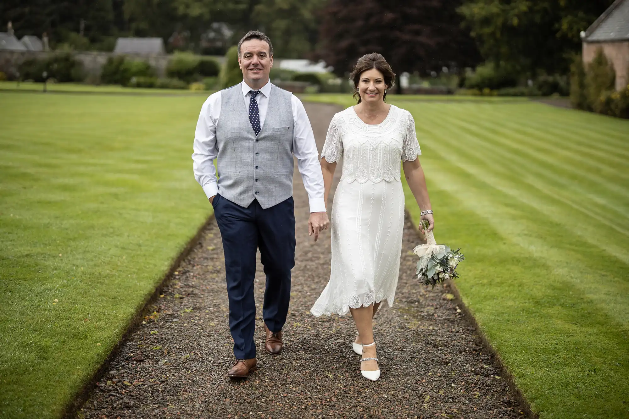 A smiling couple walk hand in hand down a gravel path on a manicured lawn. The man wears a blue suit waistcoat, tie, and white shirt; the woman wears a knee-length white lace dress and holds a bouquet. Both look happy and dressed for a wedding or formal event.